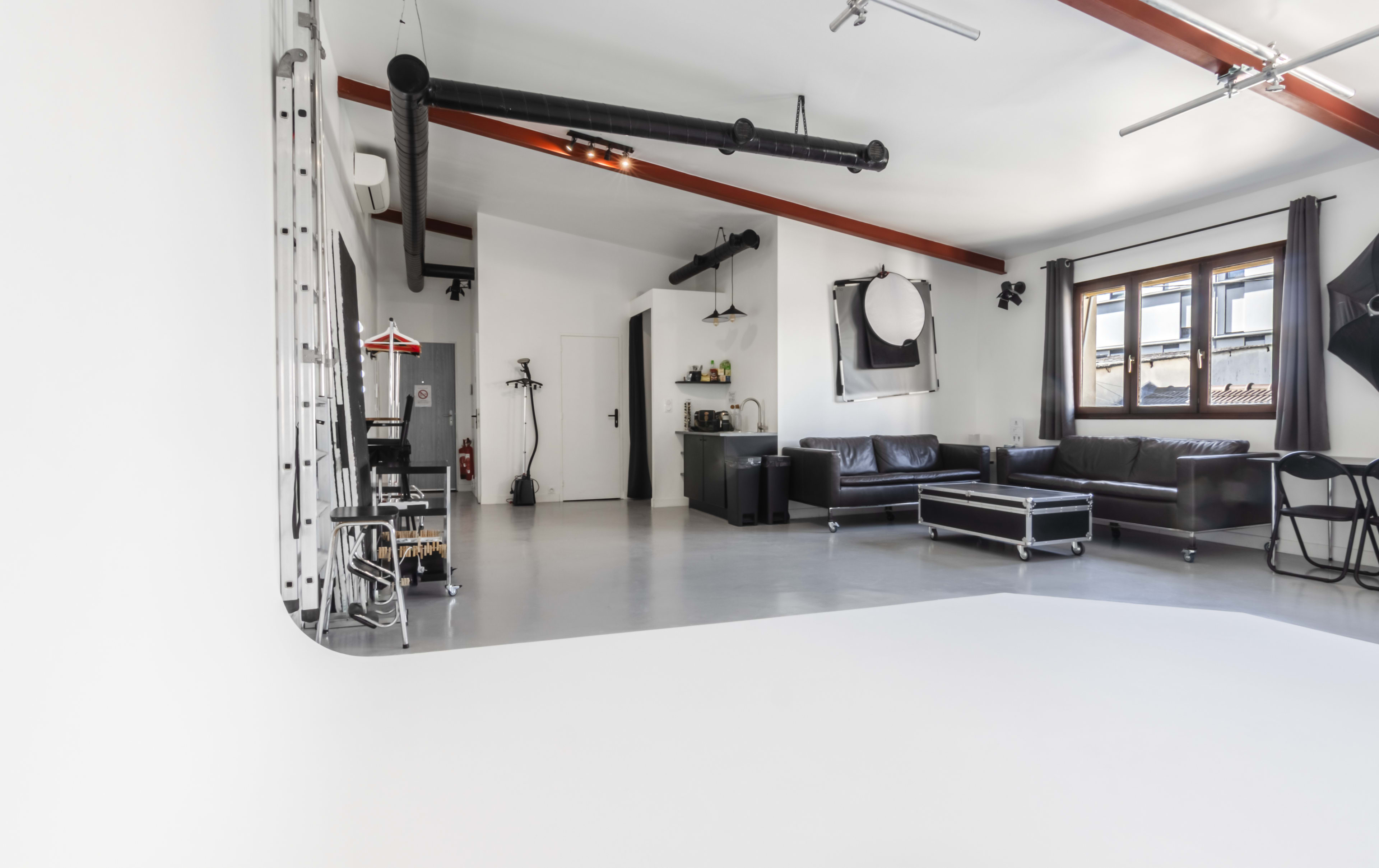 A spacious, well-lit studio with grey flooring, black furniture, and large windows.