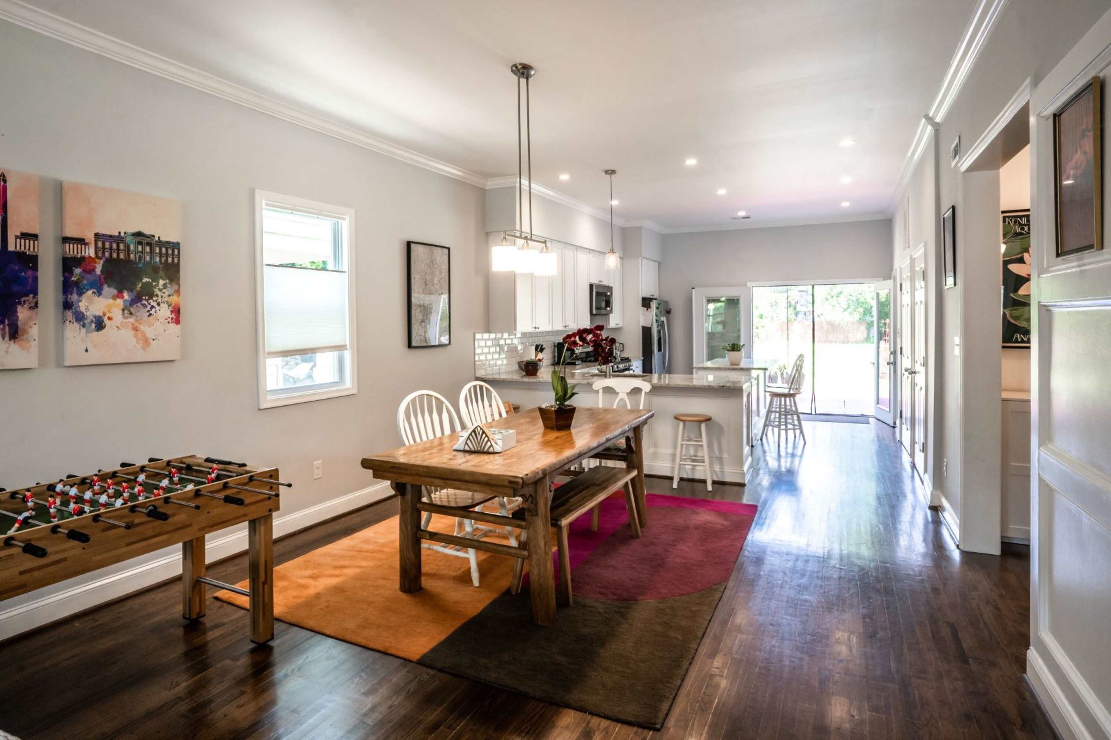 A modern open-concept kitchen and dining area with a wooden table, white chairs, and a foosball table on the side.