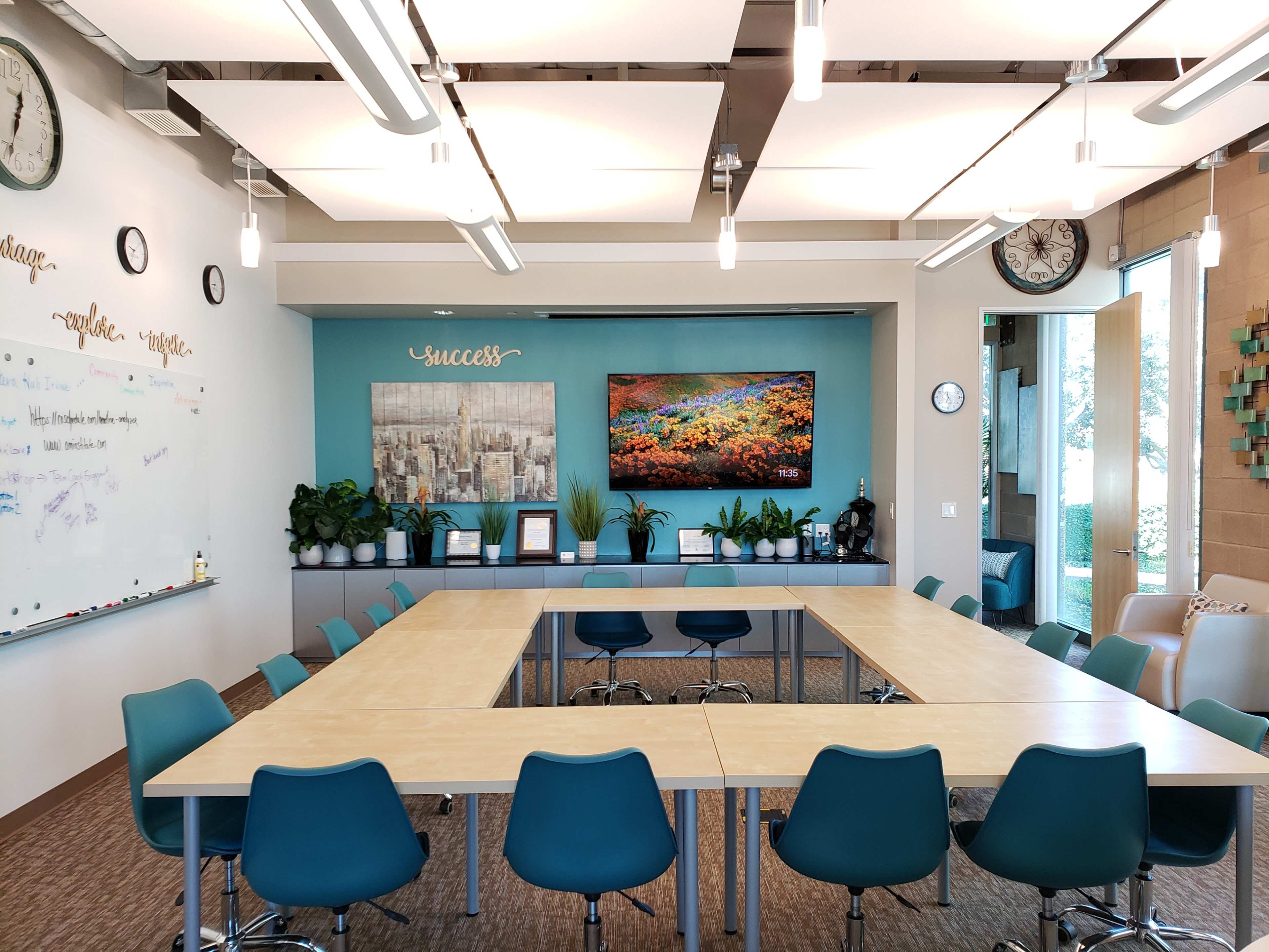 A modern conference room features a large central table surrounded by blue chairs, with plants and a wall-mounted screen displaying a landscape.