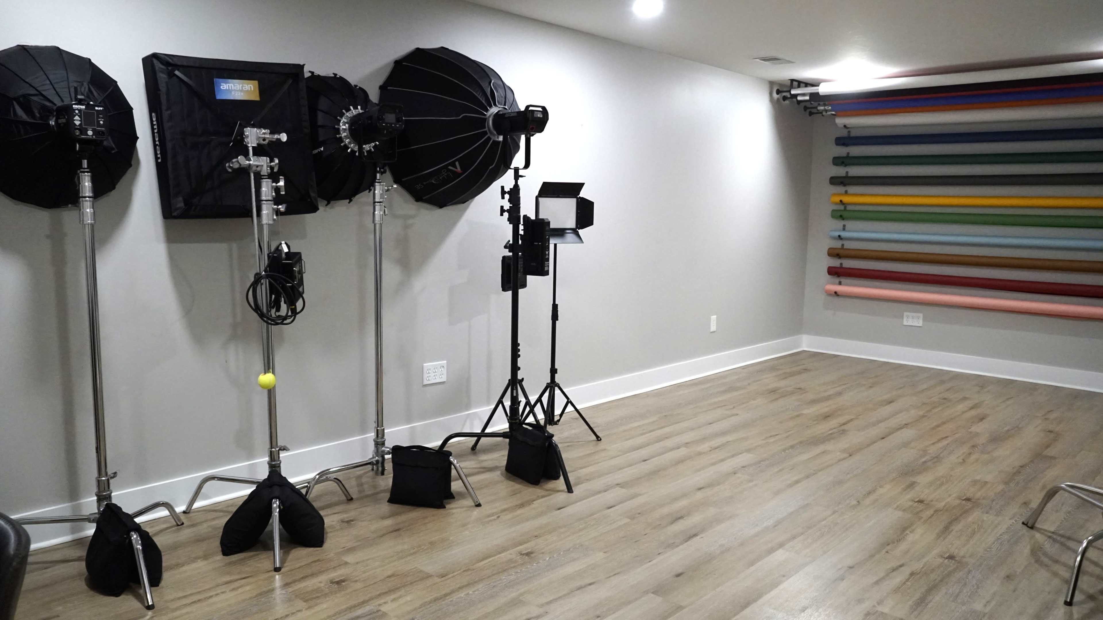 The image shows a photography studio with lighting equipment on stands and a wall lined with colorful backdrop rolls.