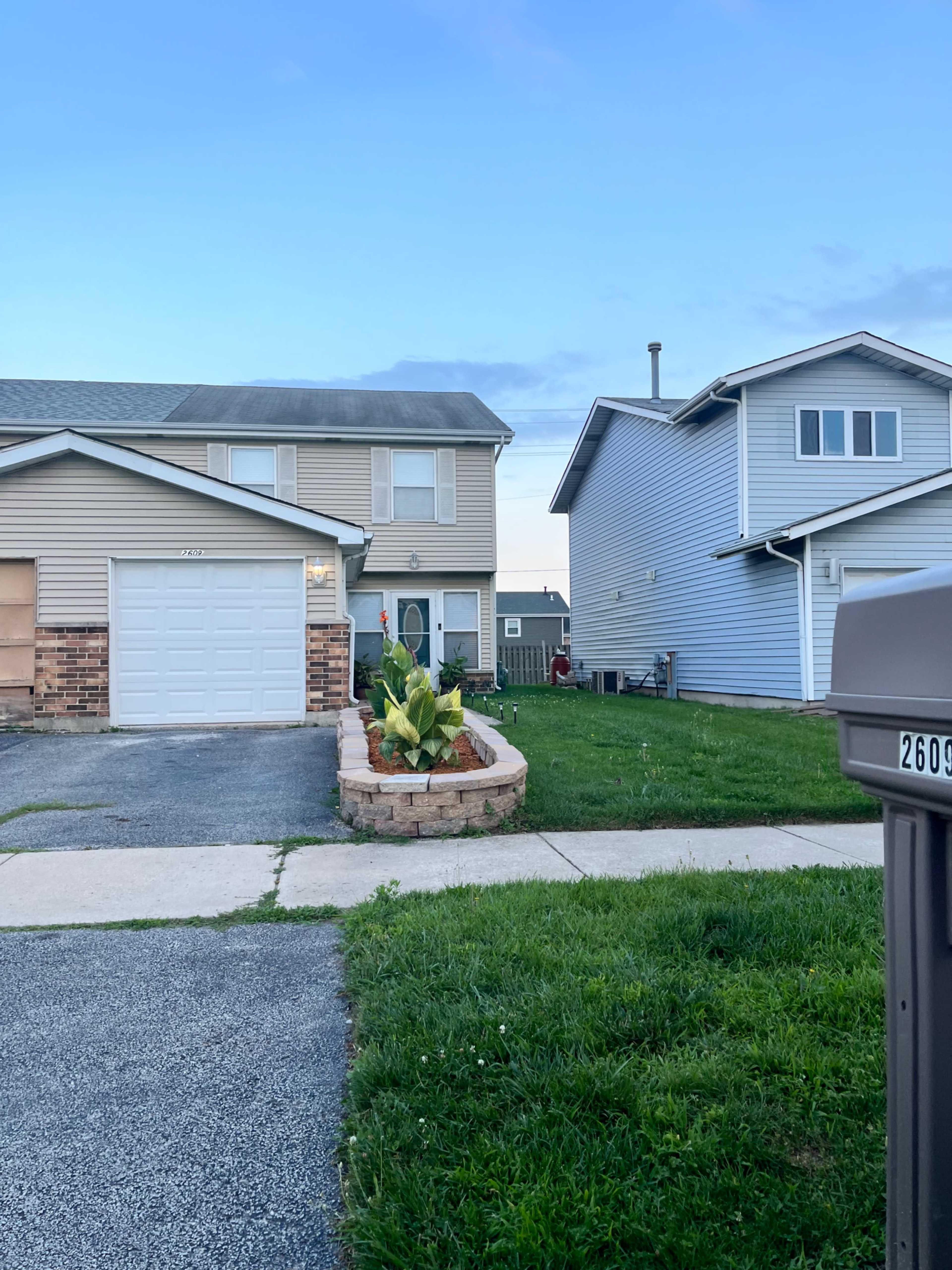 A residential area with two houses, one featuring a stone planter with plants in the front yard and a driveway leading to a garage.