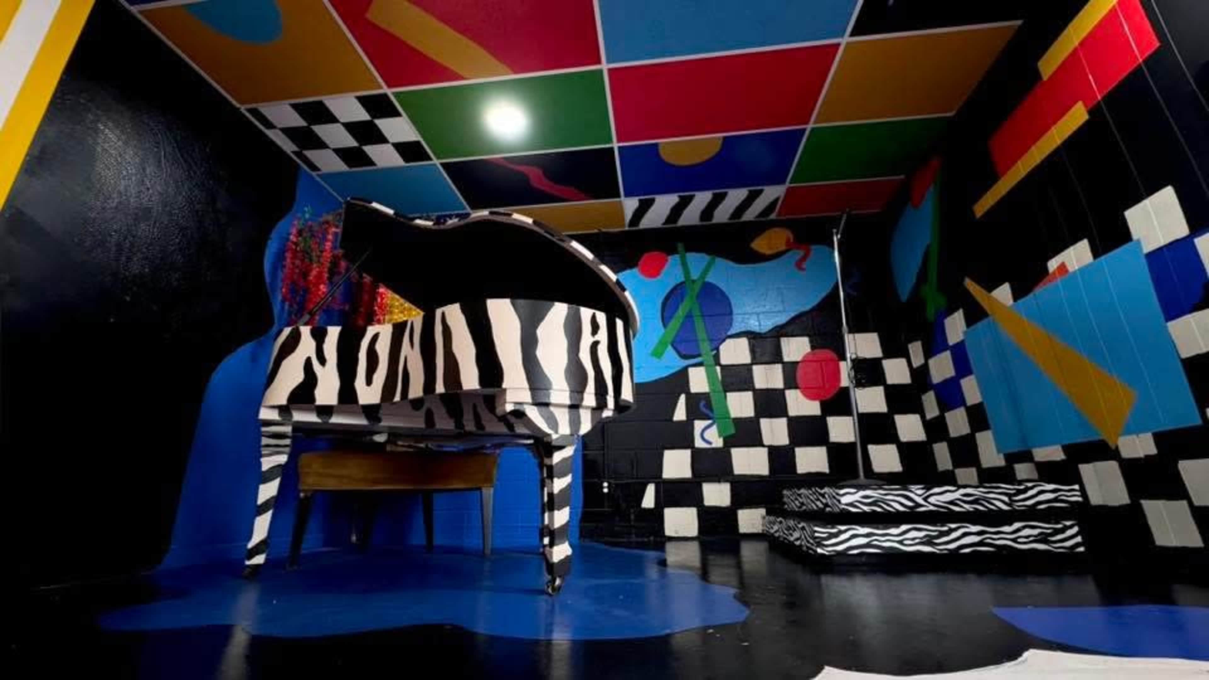 The image features a brightly colored room with abstract geometric patterns on the walls and a black-and-white striped piano in the center.