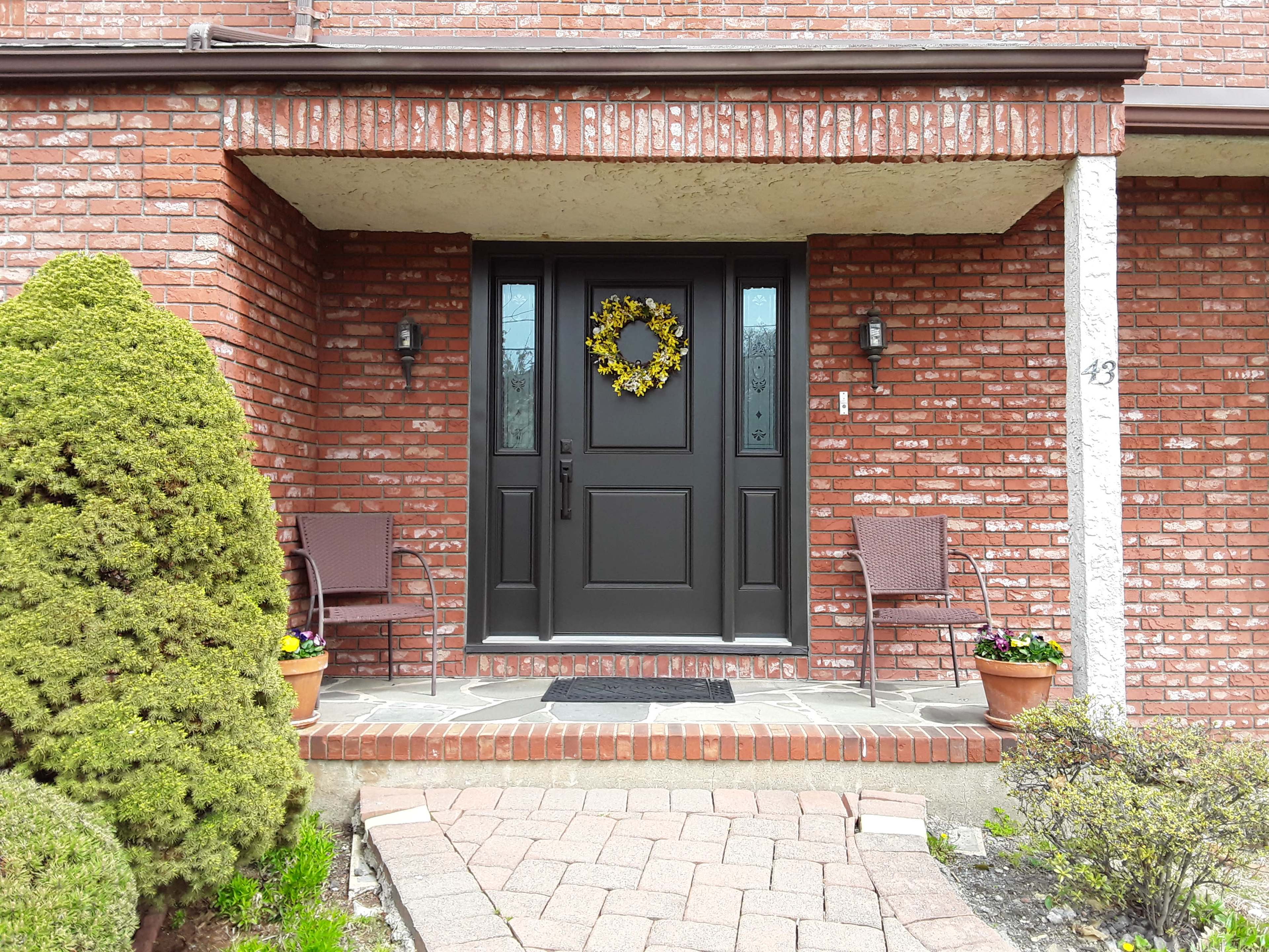 A brick house entrance featuring a dark front door adorned with a yellow wreath, flanked by two chairs and potted plants on either side.