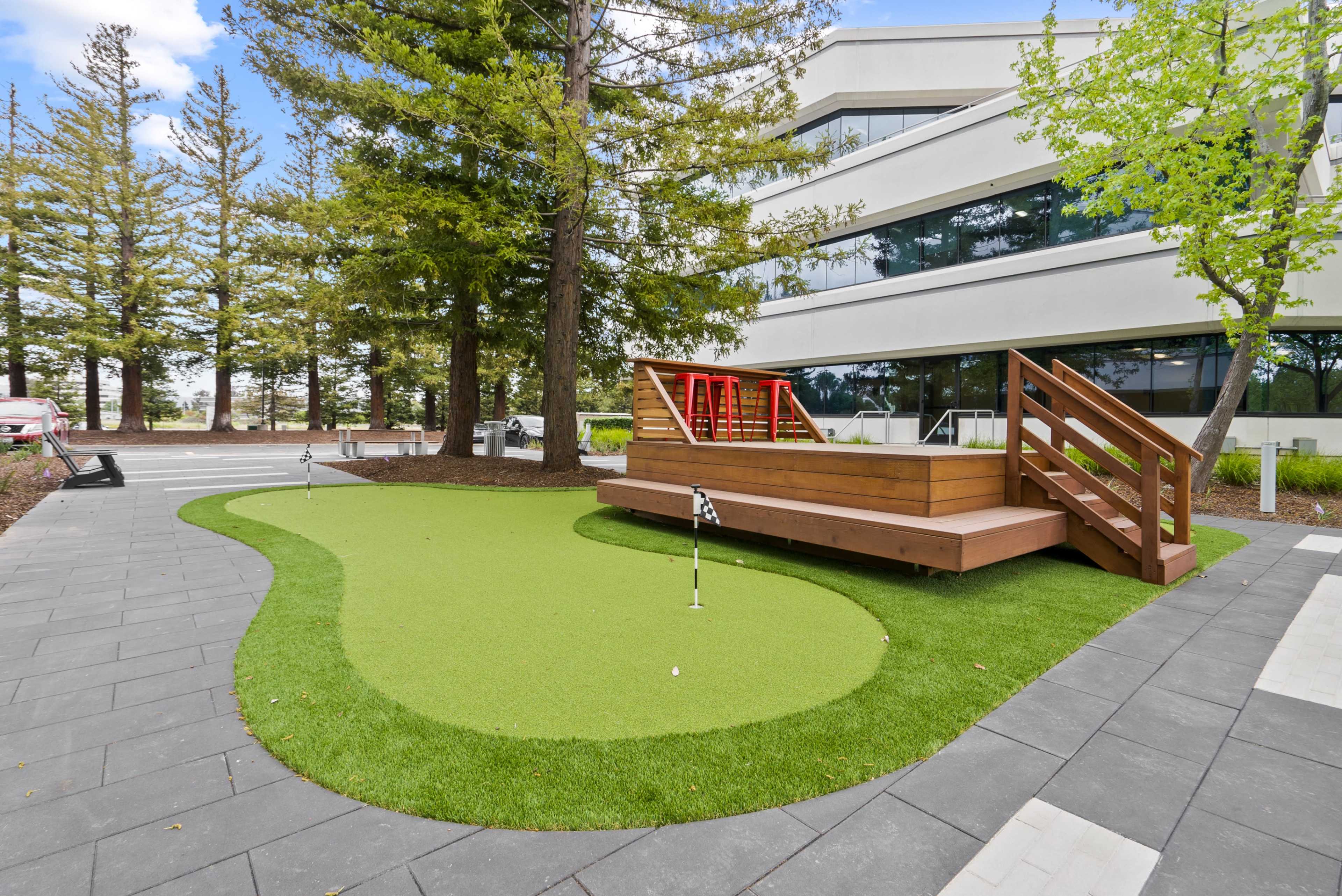 The image shows a small putting green with two holes and a wooden deck nearby, surrounded by trees and a modern building.