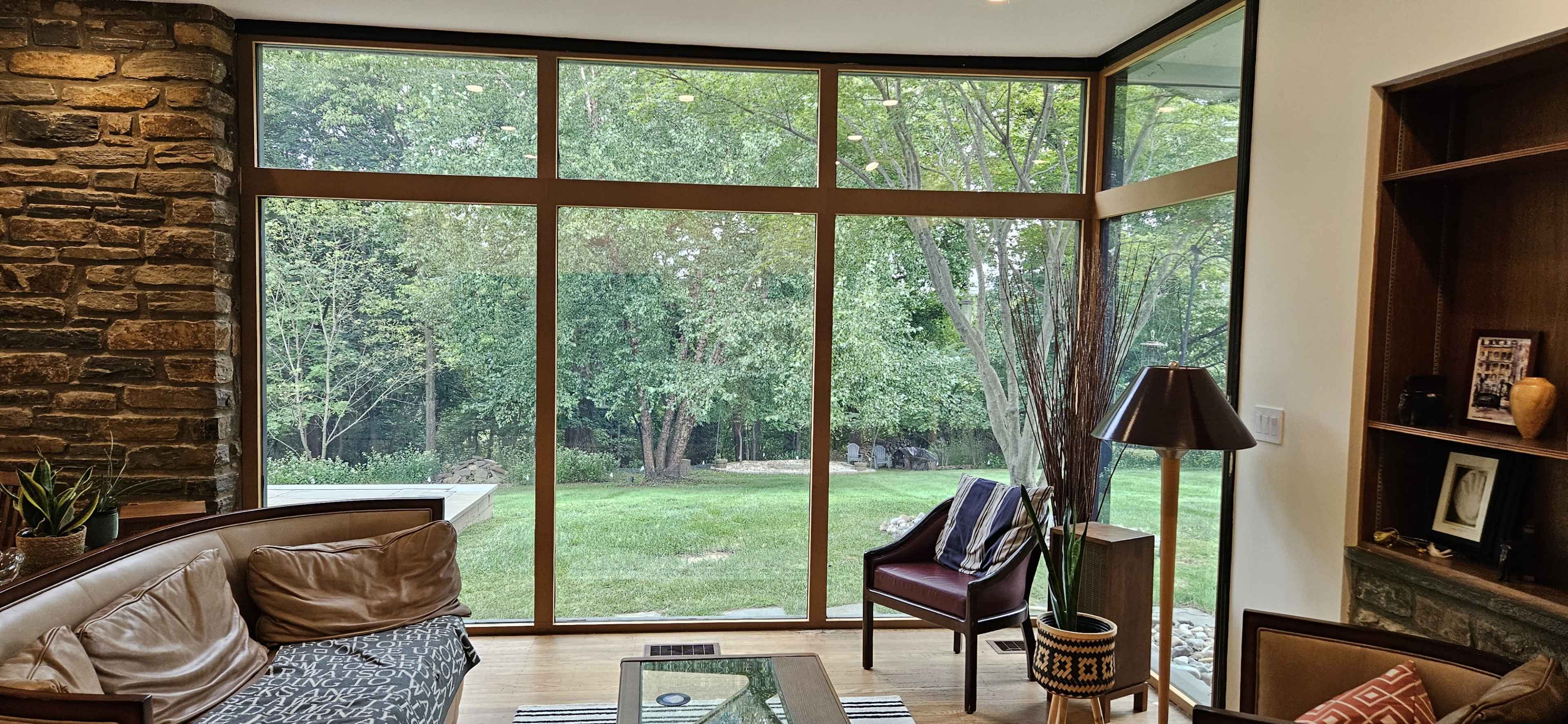 The image shows a modern living room with large windows that offer a view of a green garden filled with trees.
