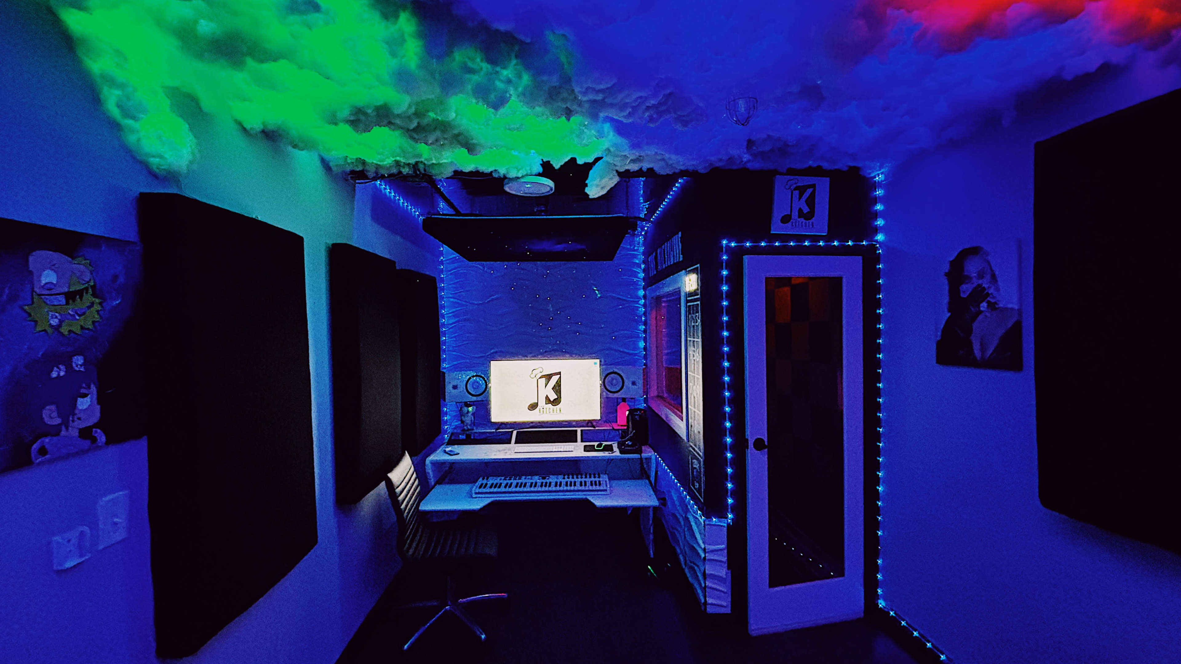 The image depicts a modern music studio with colorful LED lighting, soundproofing panels, a computer workstation, and a door leading to another room.