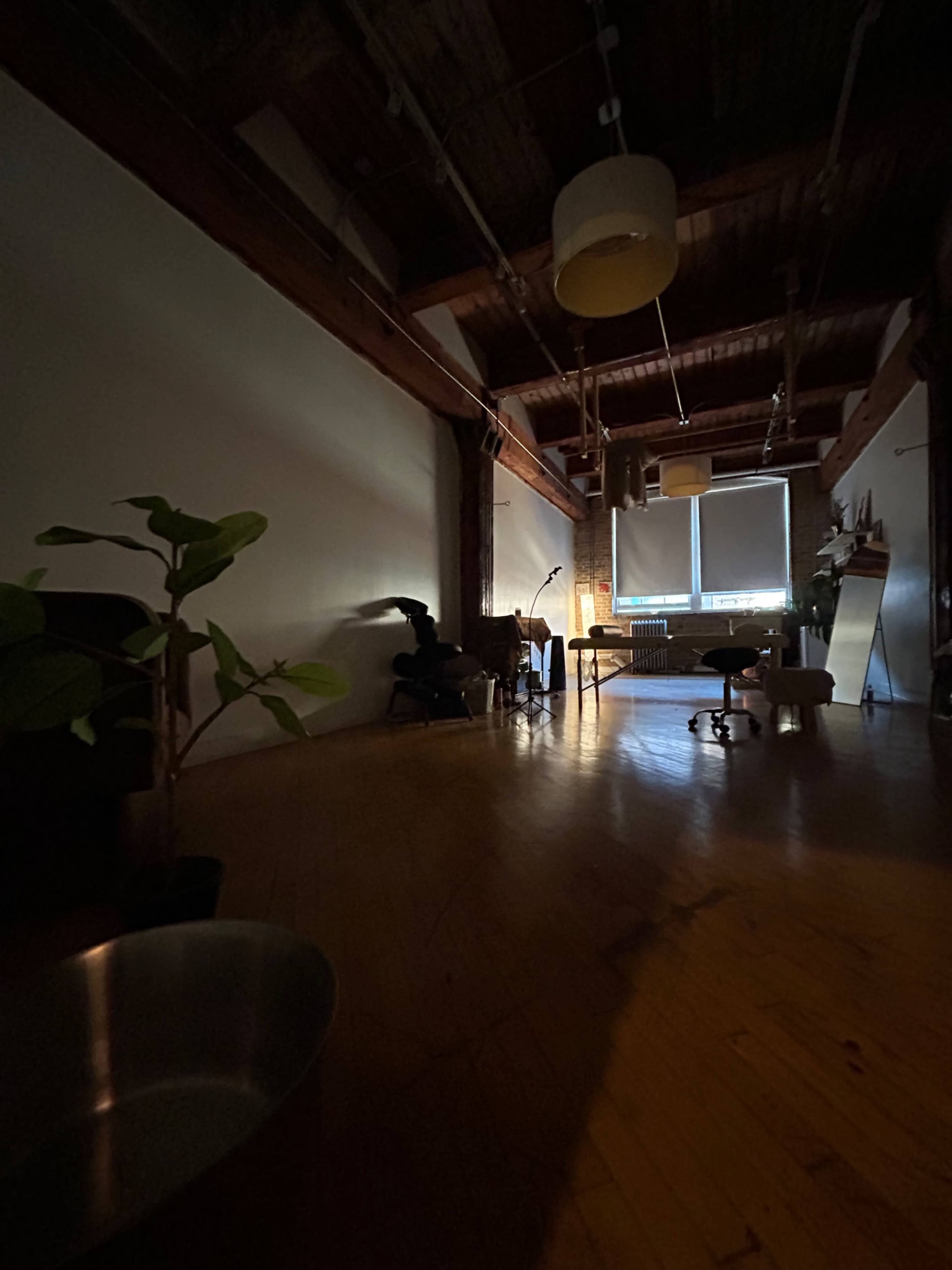 The image shows a dimly lit room with wooden floors, a few chairs, and a large window letting in natural light.