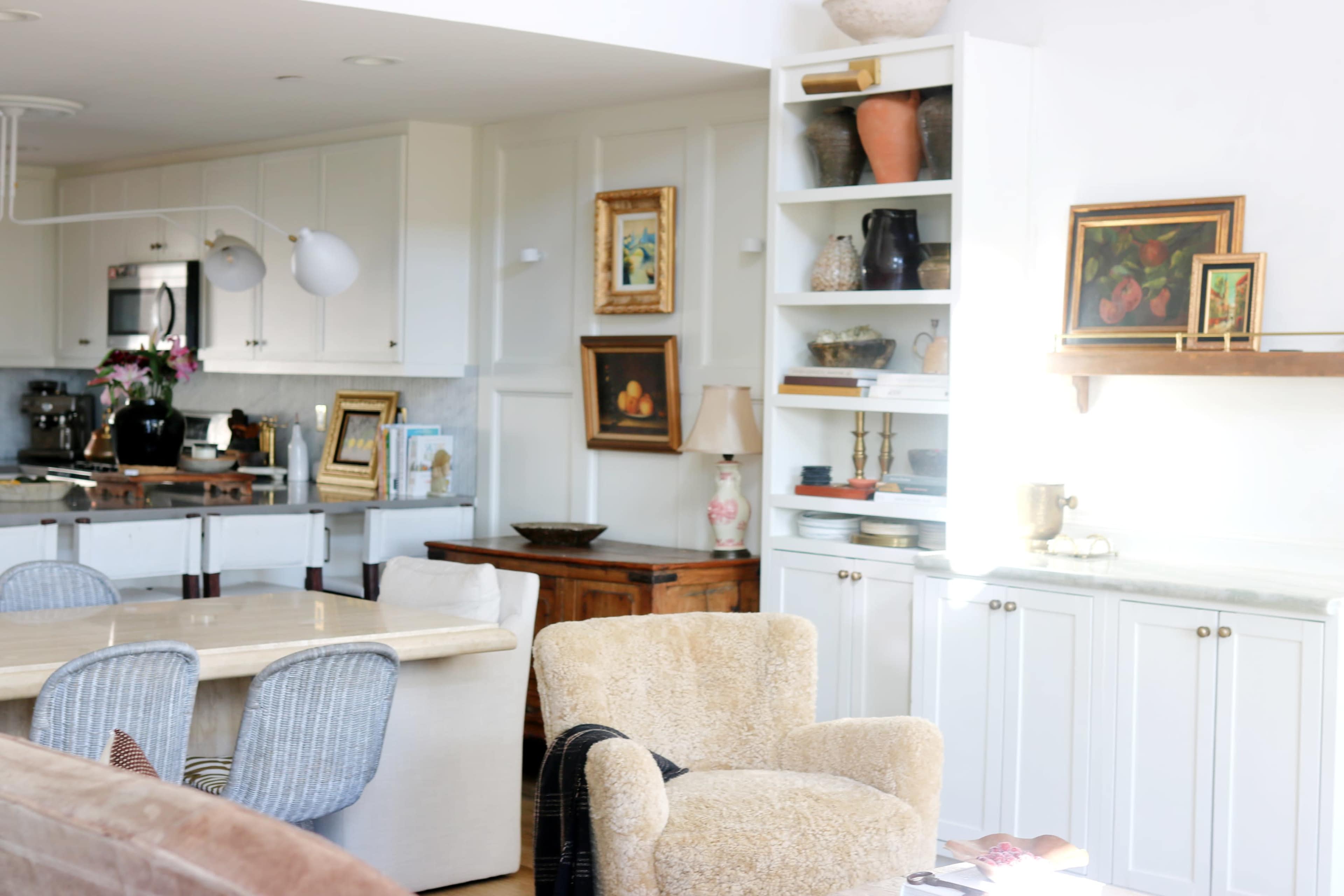 A modern kitchen and dining area features a light-colored marble table, woven chairs, and shelves displaying various decorative items.