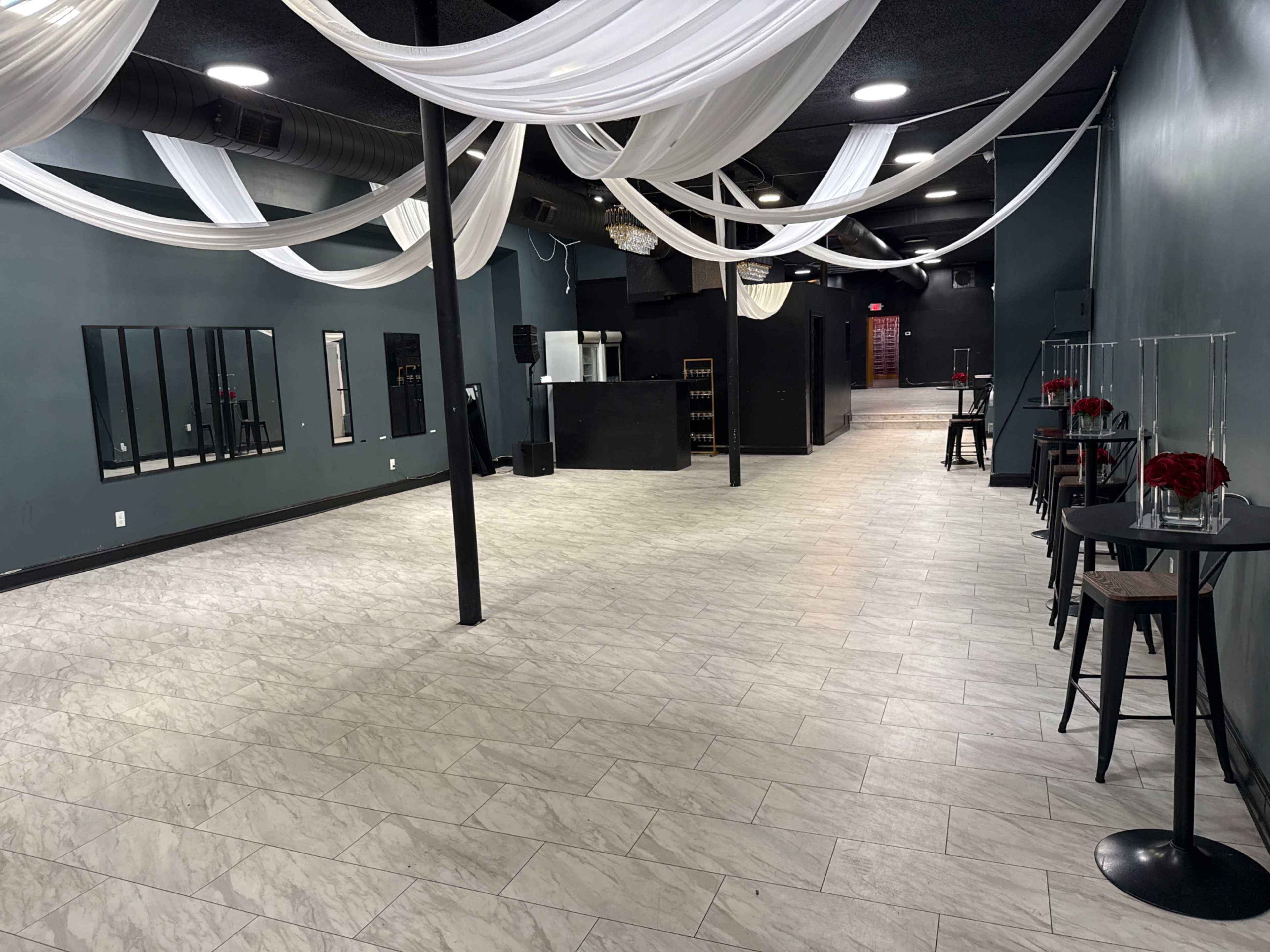 The image shows an empty event space with a black bar area, decorative drapes hanging from the ceiling, and small tables along the walls.