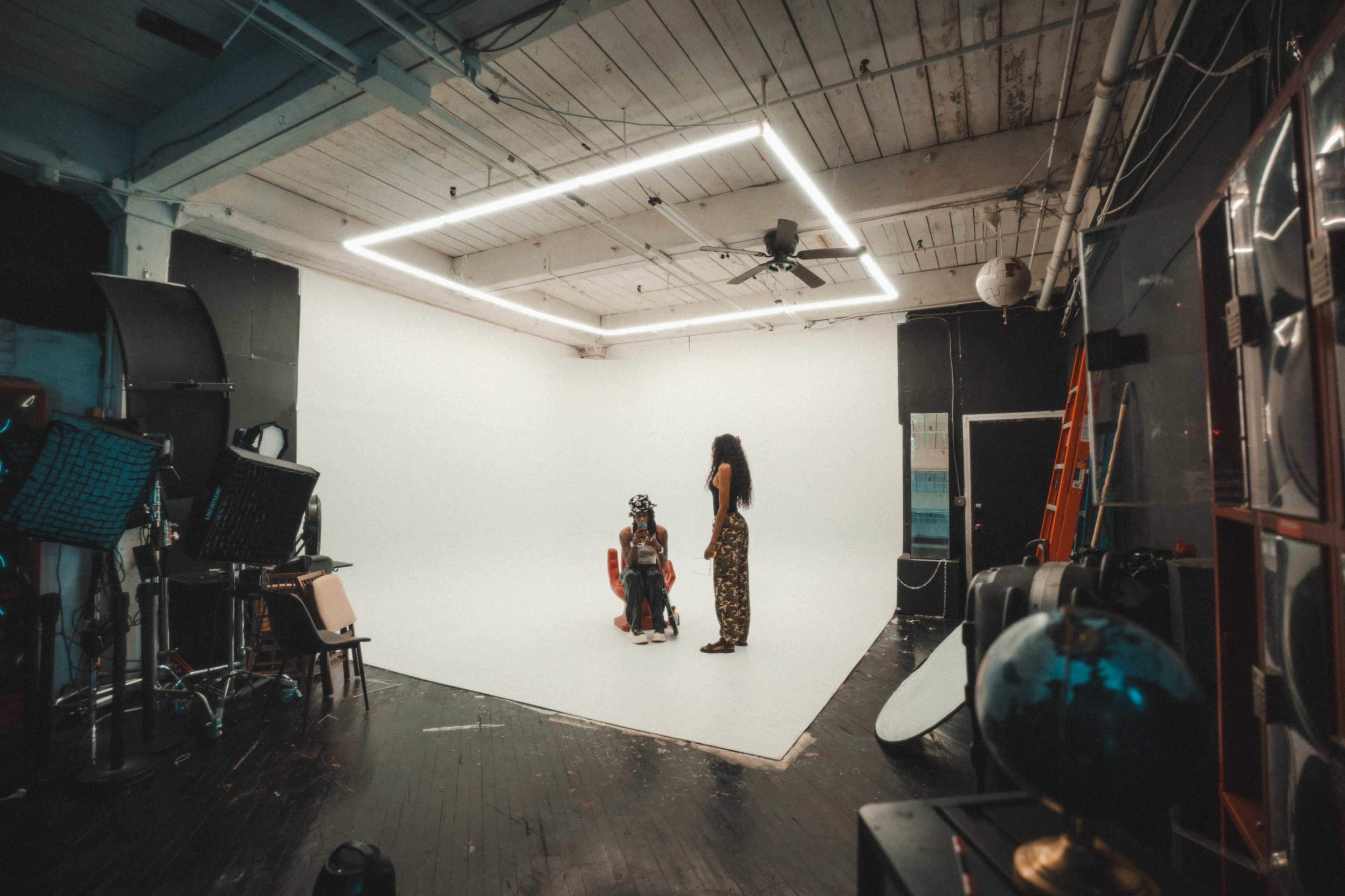 A brightly lit photography studio features a white backdrop with two individuals interacting on the floor and various equipment lined along the walls.