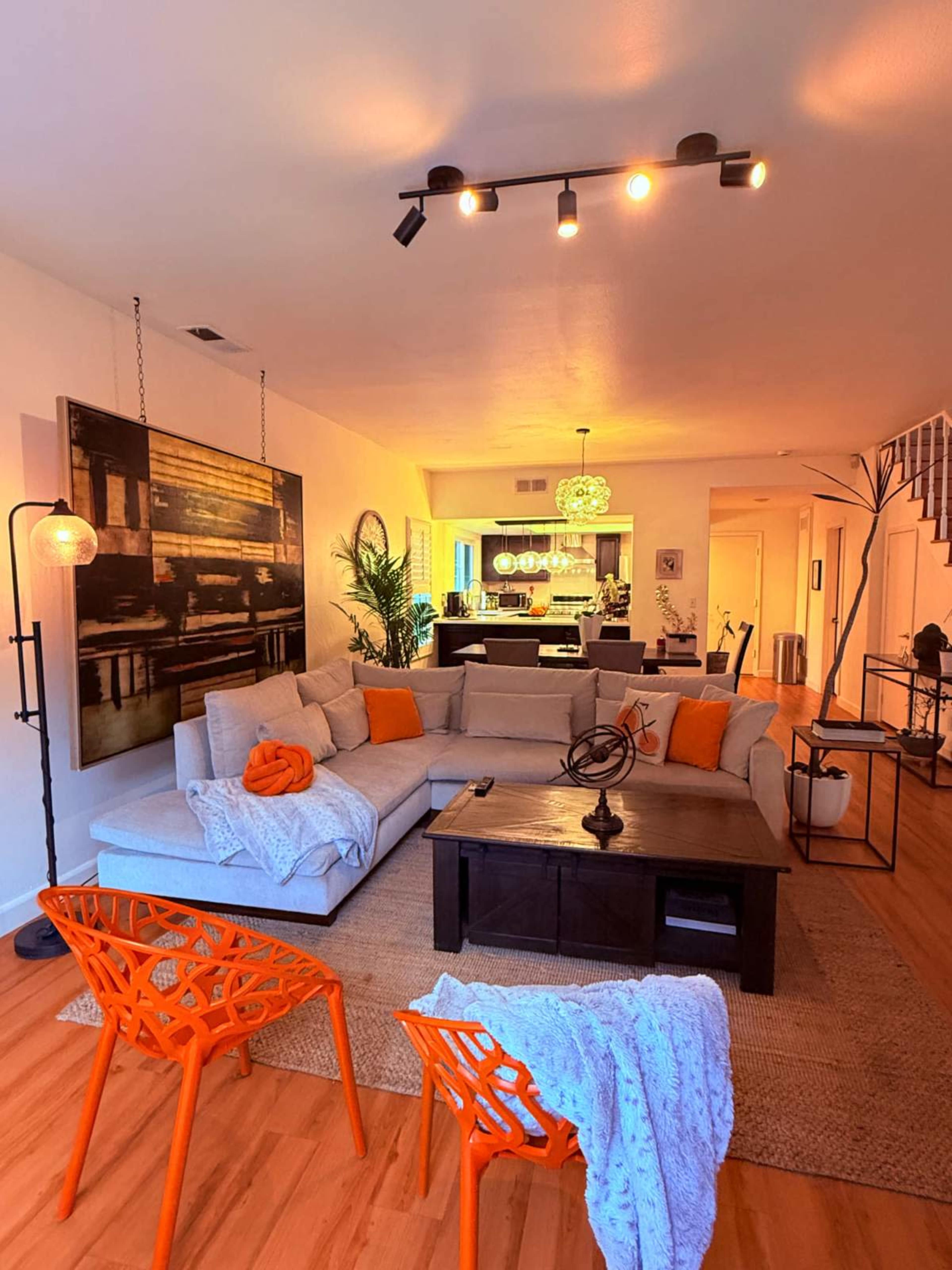 The image shows a modern living room with a light gray sectional sofa, orange accents, a wooden coffee table, and orange chairs, leading to a kitchen area in the background.