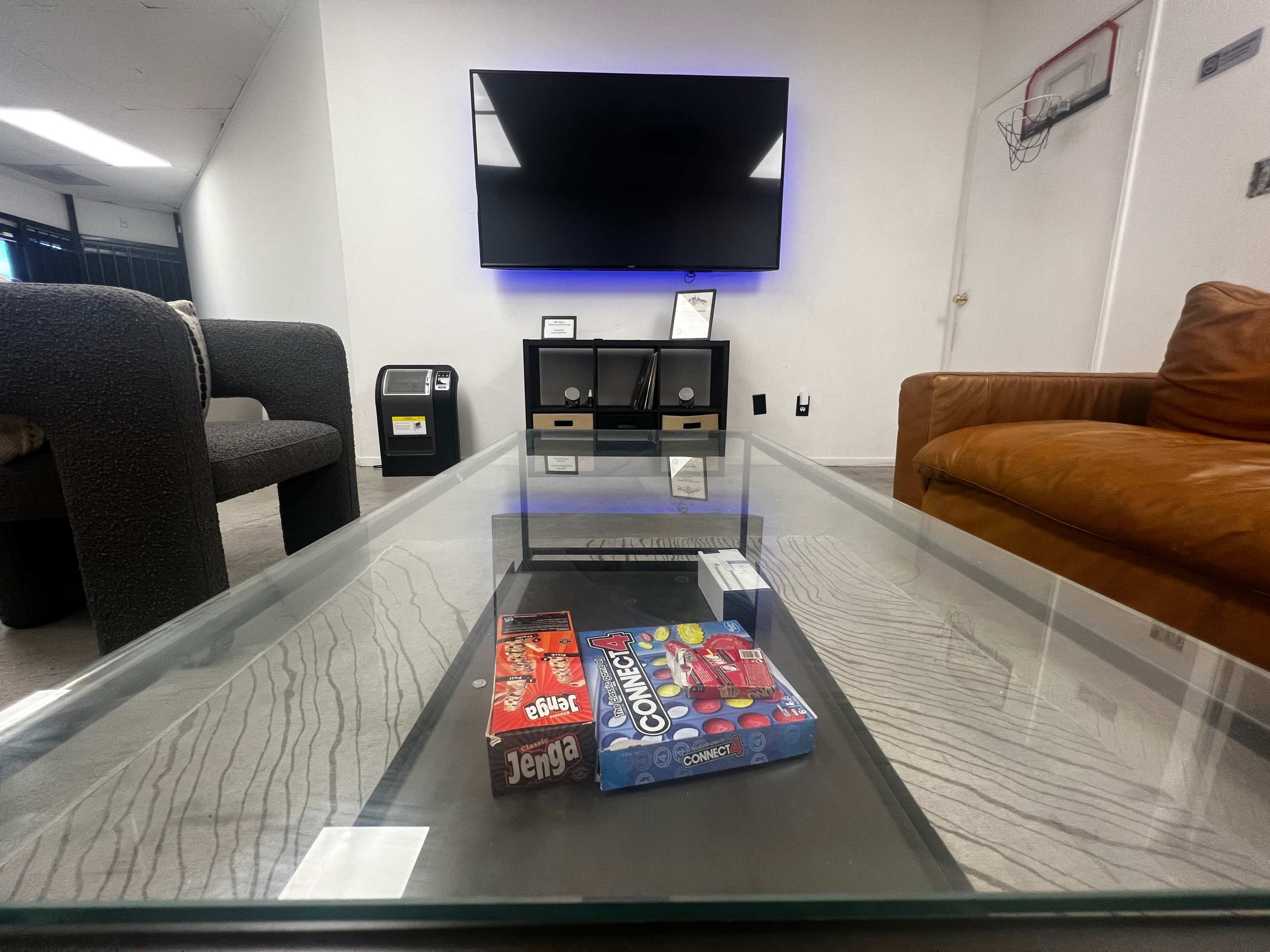 The scene features a modern living room with a glass coffee table holding board games, a large wall-mounted TV, and a brown leather couch.