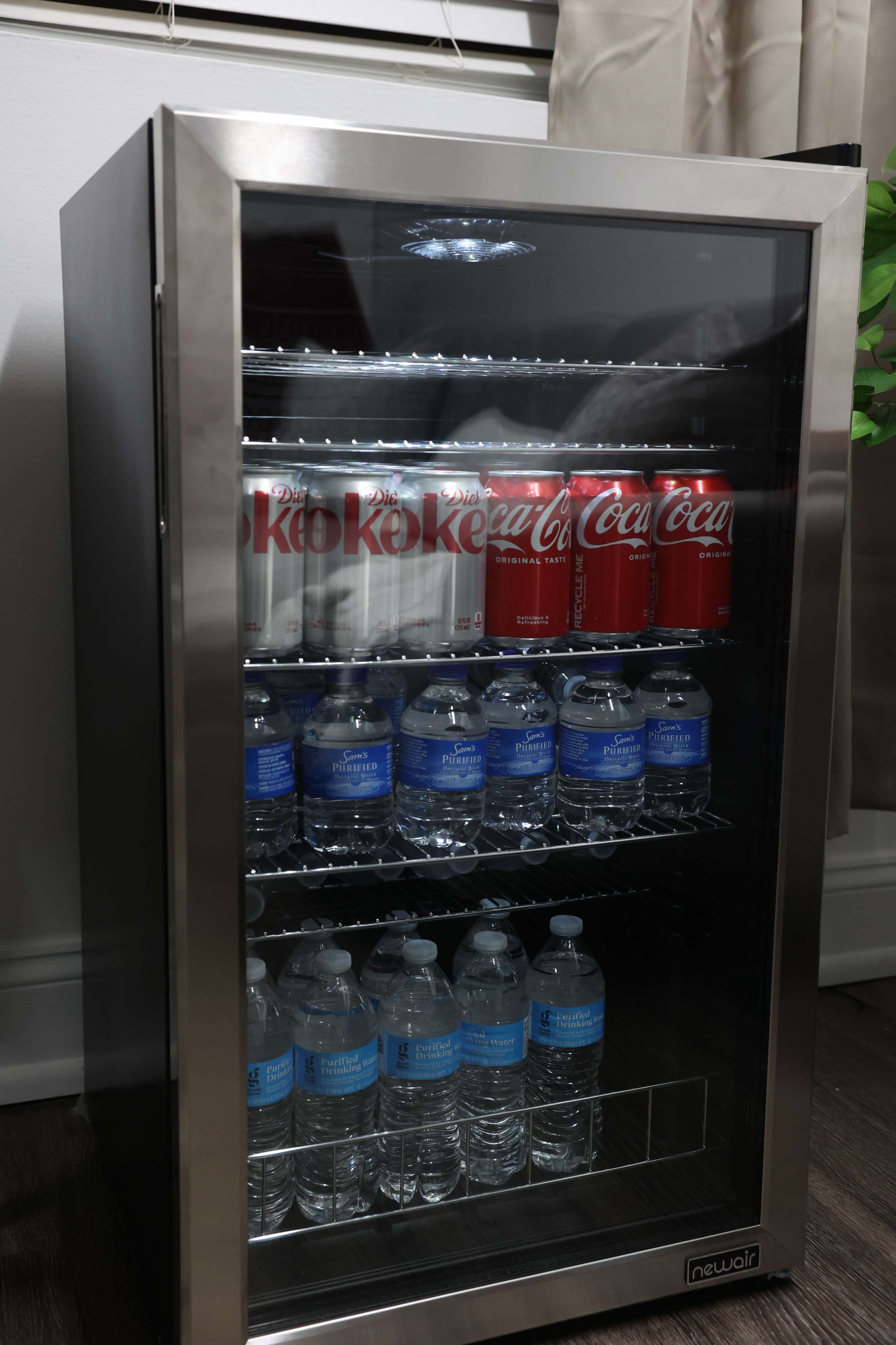 The image shows a glass-fronted beverage refrigerator containing cans of Diet Coke and Coca-Cola on the top shelf and bottles of water on the bottom shelf.