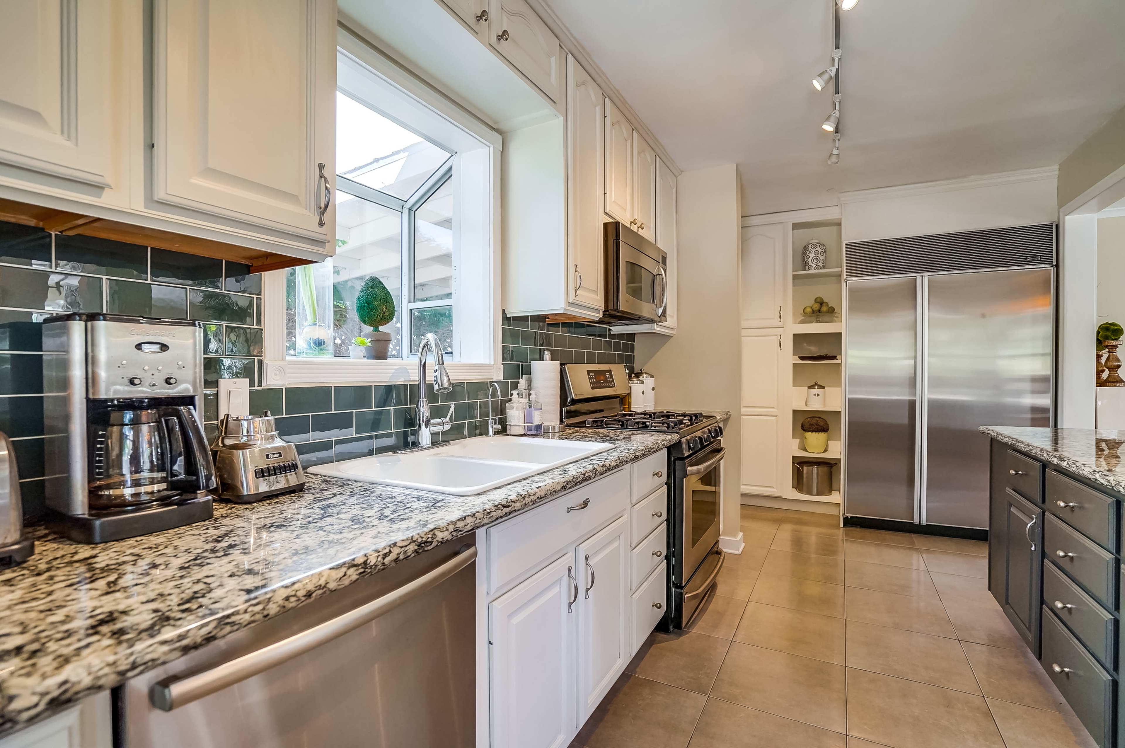 A modern kitchen features white cabinetry, a granite countertop, stainless steel appliances, and a window with a garden view.