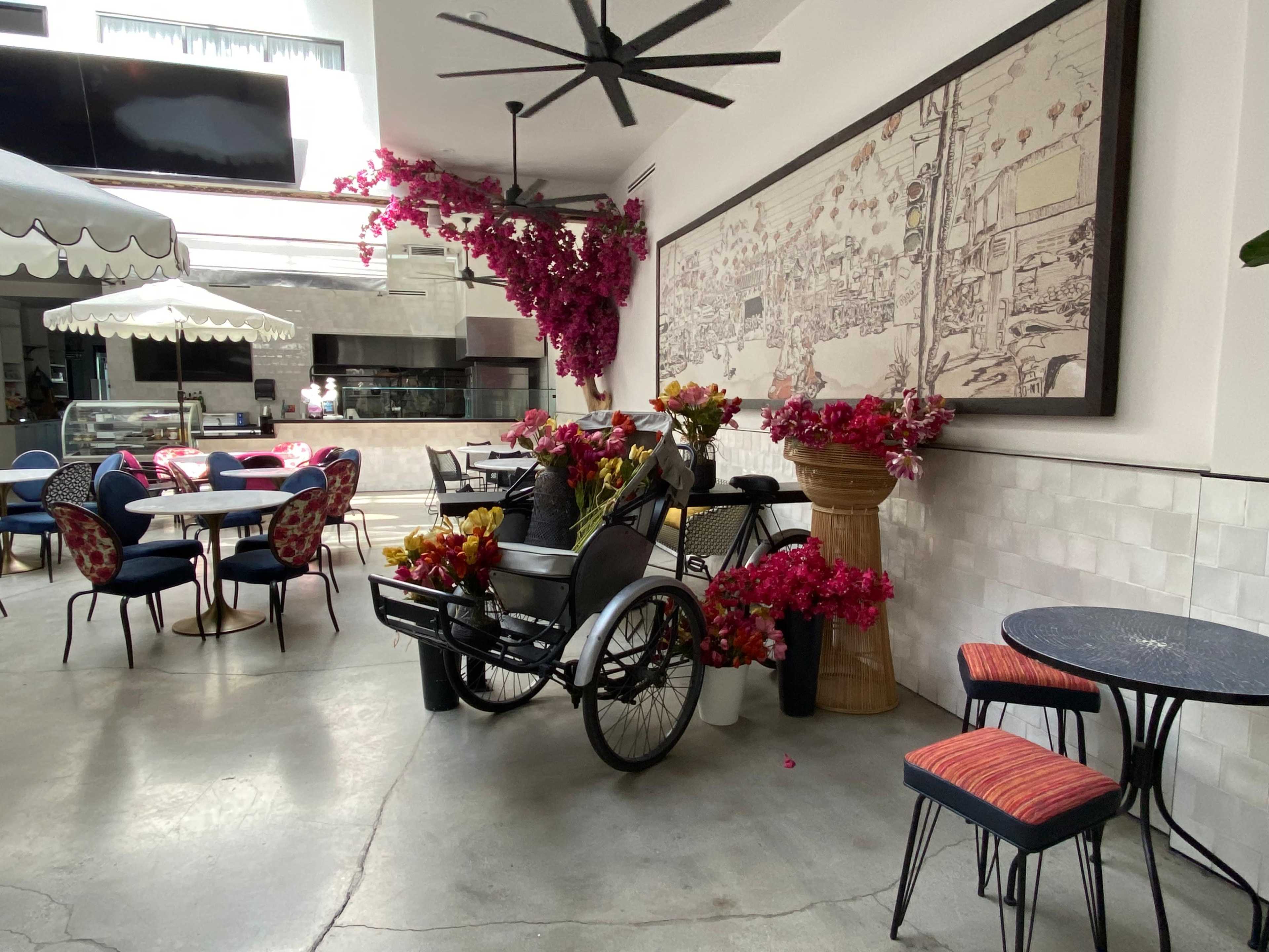 The image shows a stylish indoor café with a vintage bicycle cart adorned with flowers, surrounded by seating arrangements and decorative artwork on the walls.