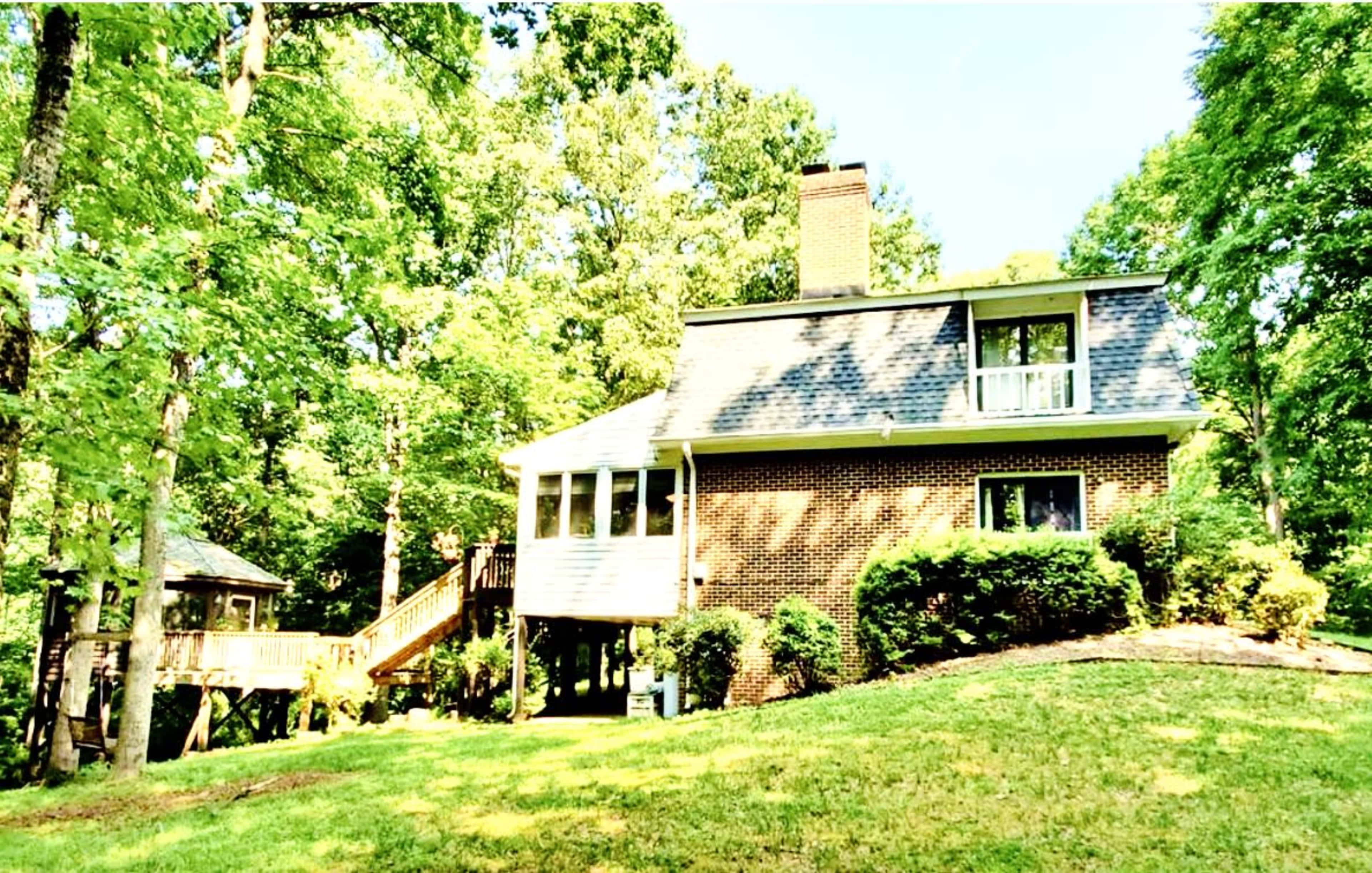A two-story brick house is surrounded by trees with a deck and staircase leading to the backyard.