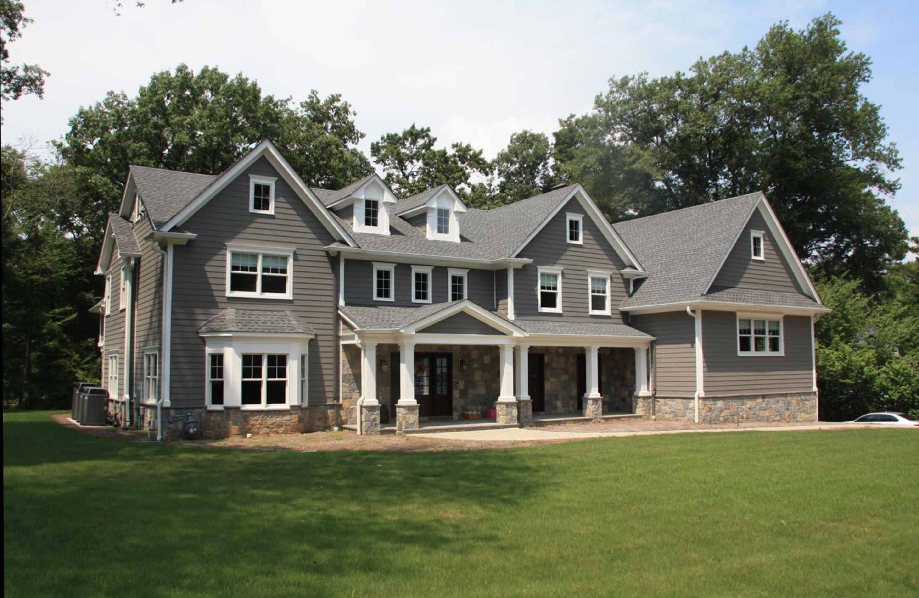 A large gray multi-story house with a stone foundation and multiple gabled roofs is set on a grassy lawn surrounded by trees.