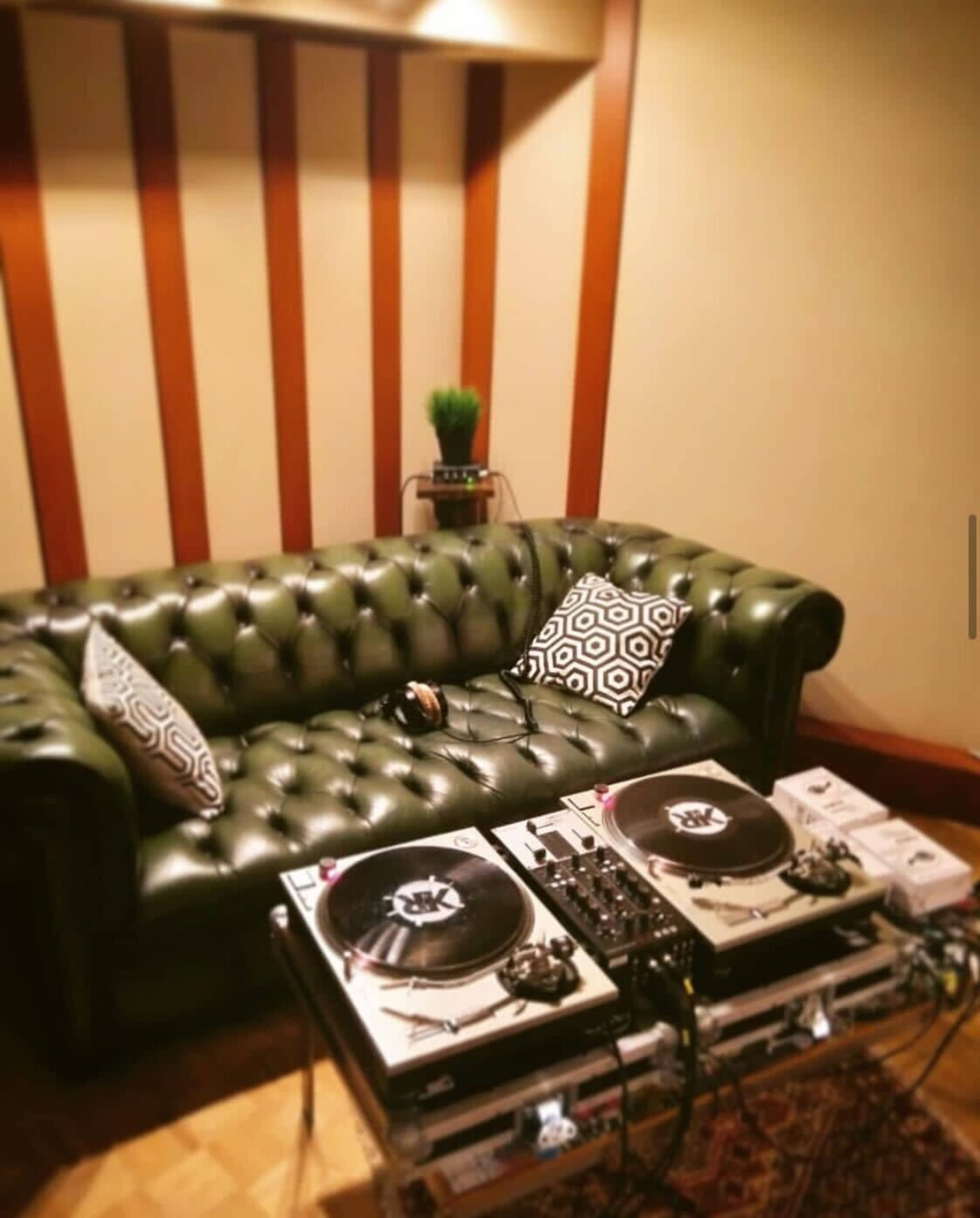 A green leather chesterfield sofa next to a pair of turntables set on a glass table in a room with wooden wall panels.