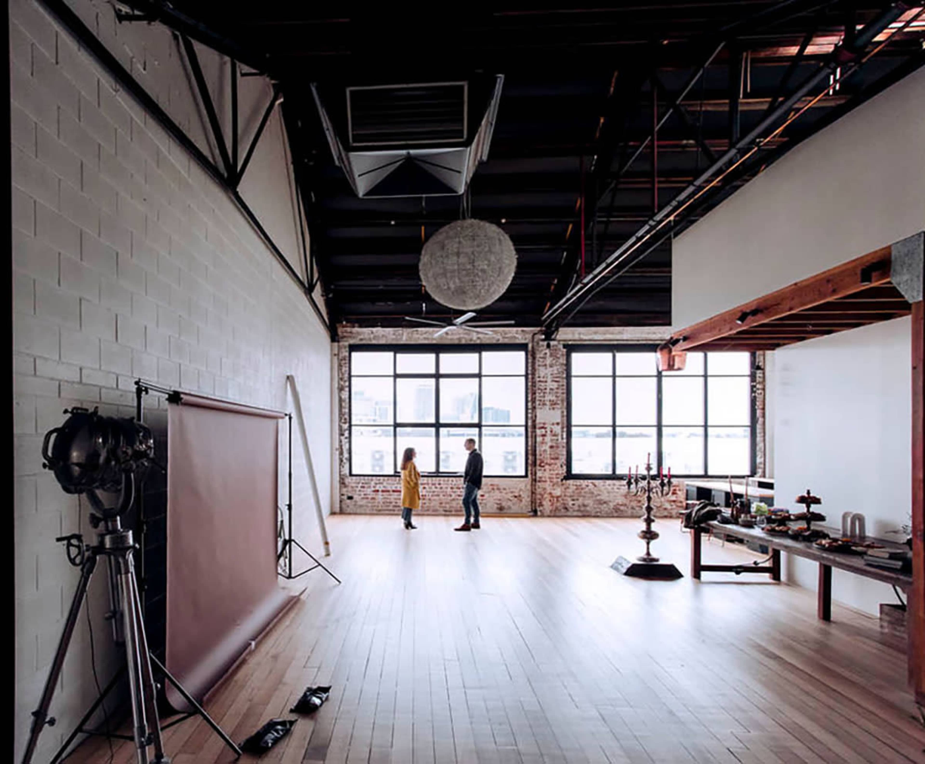 A spacious industrial studio with large windows, a smooth wooden floor, and a few scattered props, including a backdrop, lighting equipment, and a table with various items.