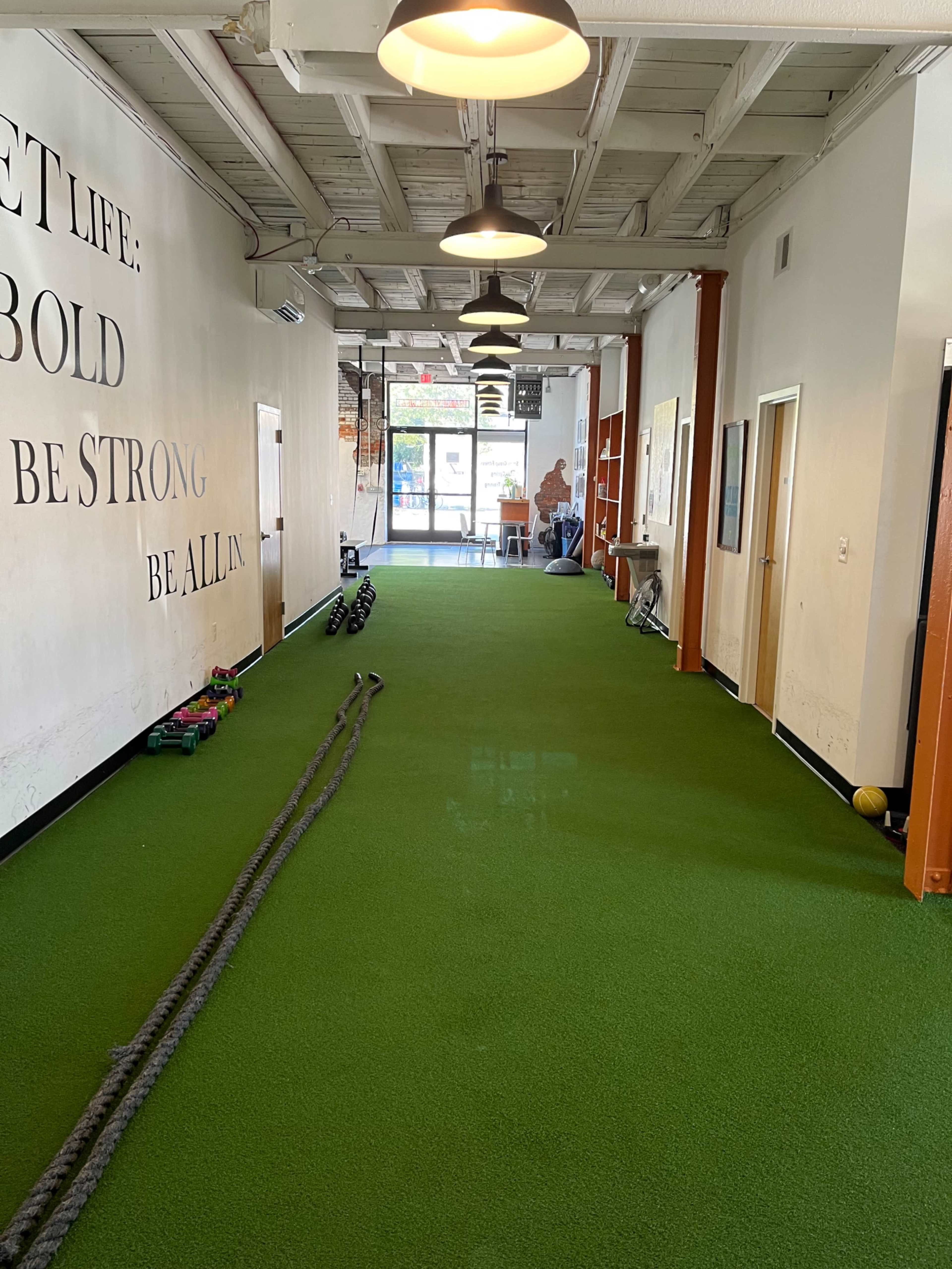 A long hallway with green turf flooring features workout equipment such as ropes and weights along the sides, leading to a glass entrance at the far end.
