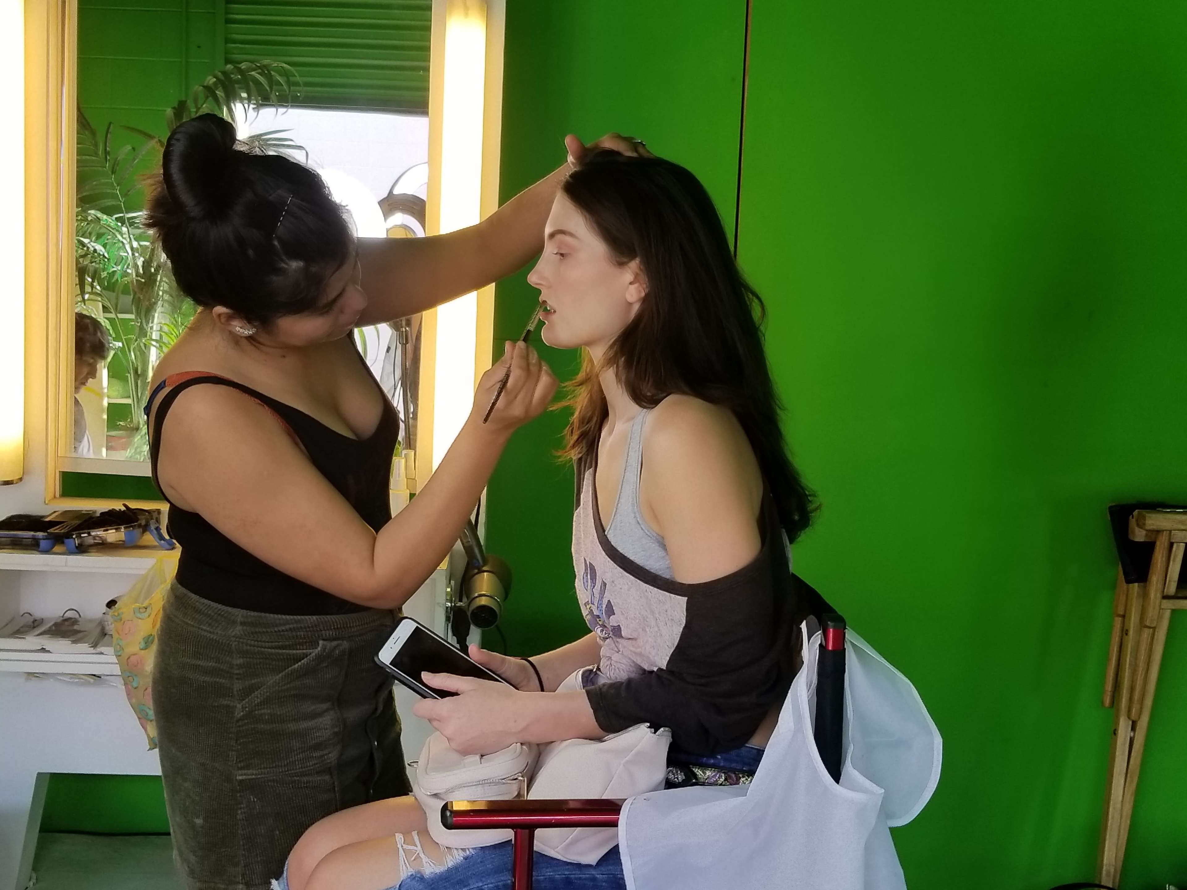 A makeup artist applies makeup to a woman sitting in a chair in front of a green backdrop.