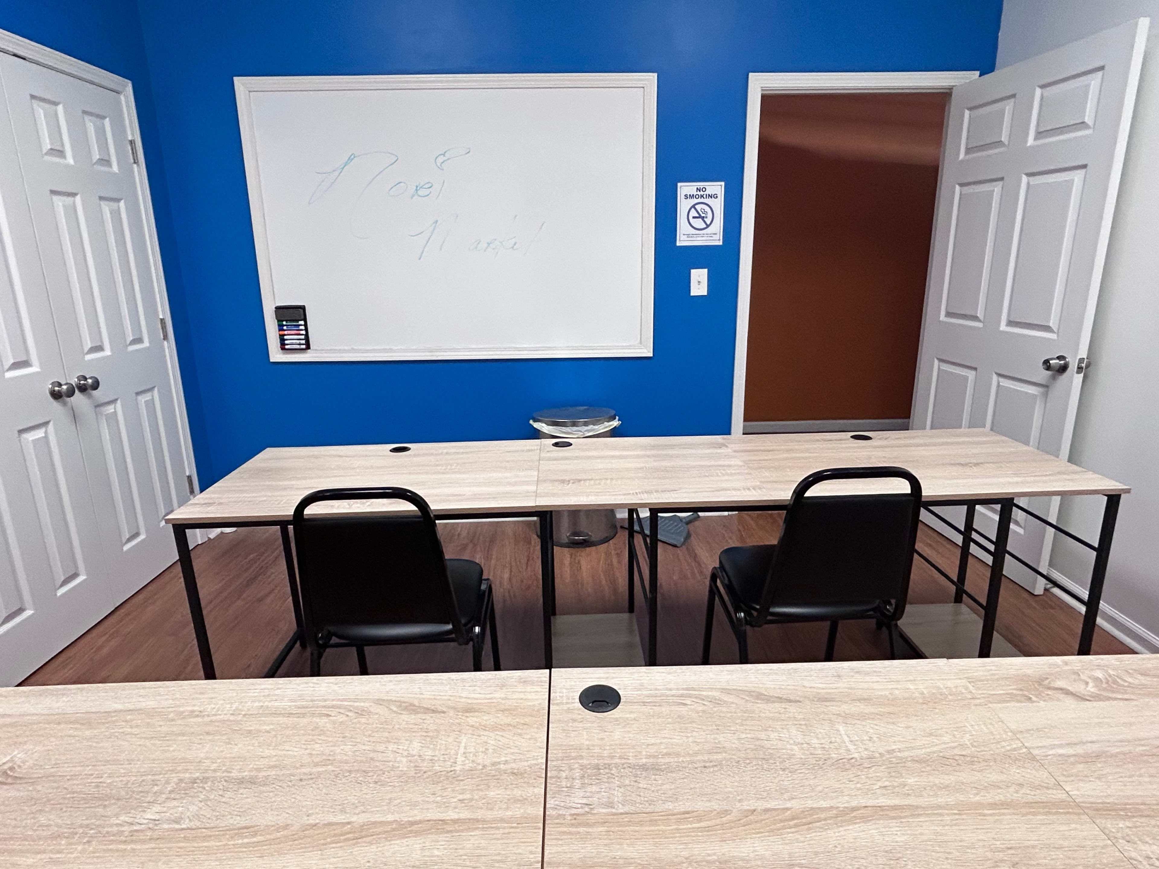 The image shows a classroom with two empty chairs and a long table facing a whiteboard on a blue wall.