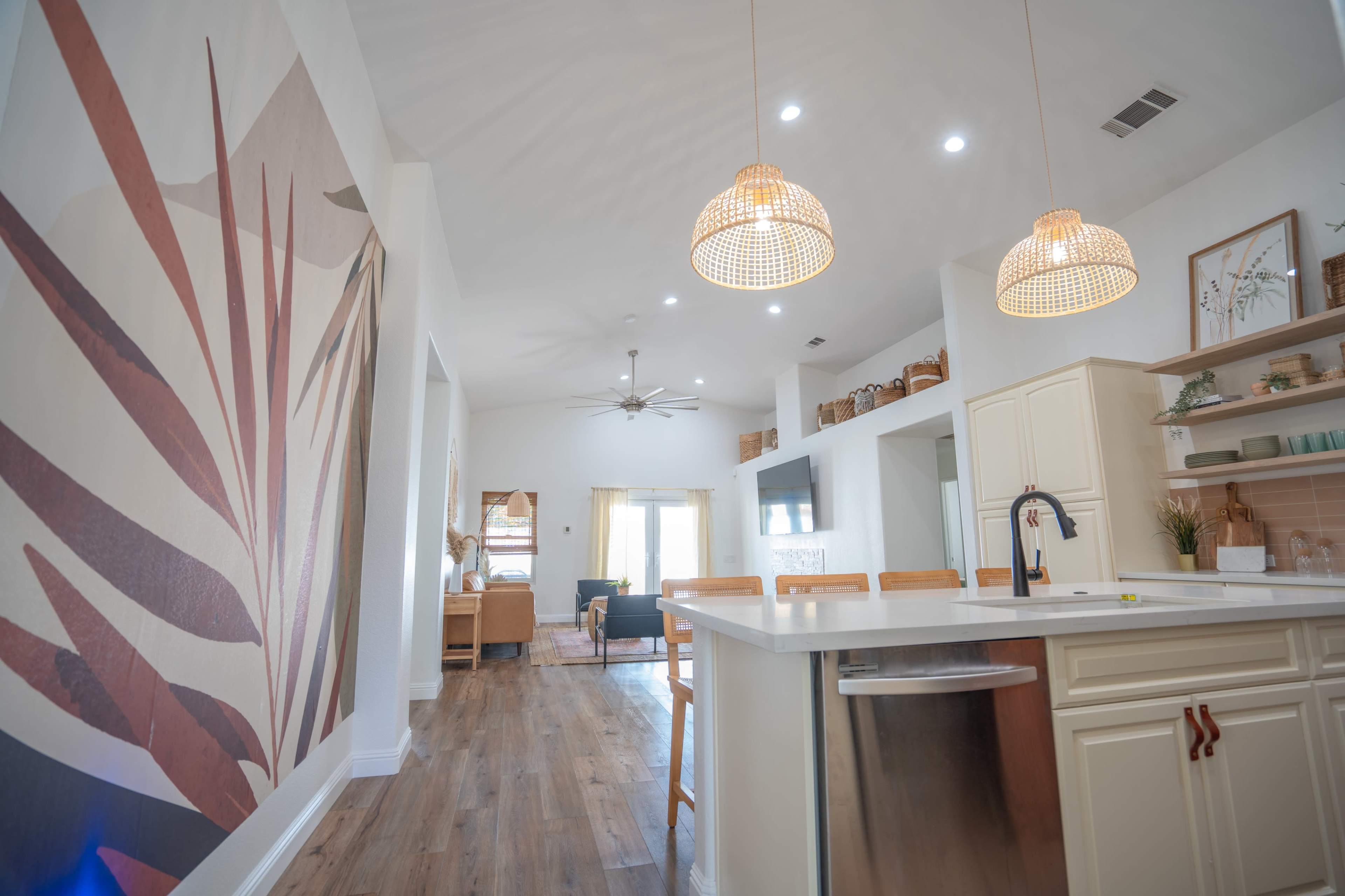 A modern kitchen with a white island, pendant lights, and a large mural featuring stylized plant leaves, leading into a living space with natural light.