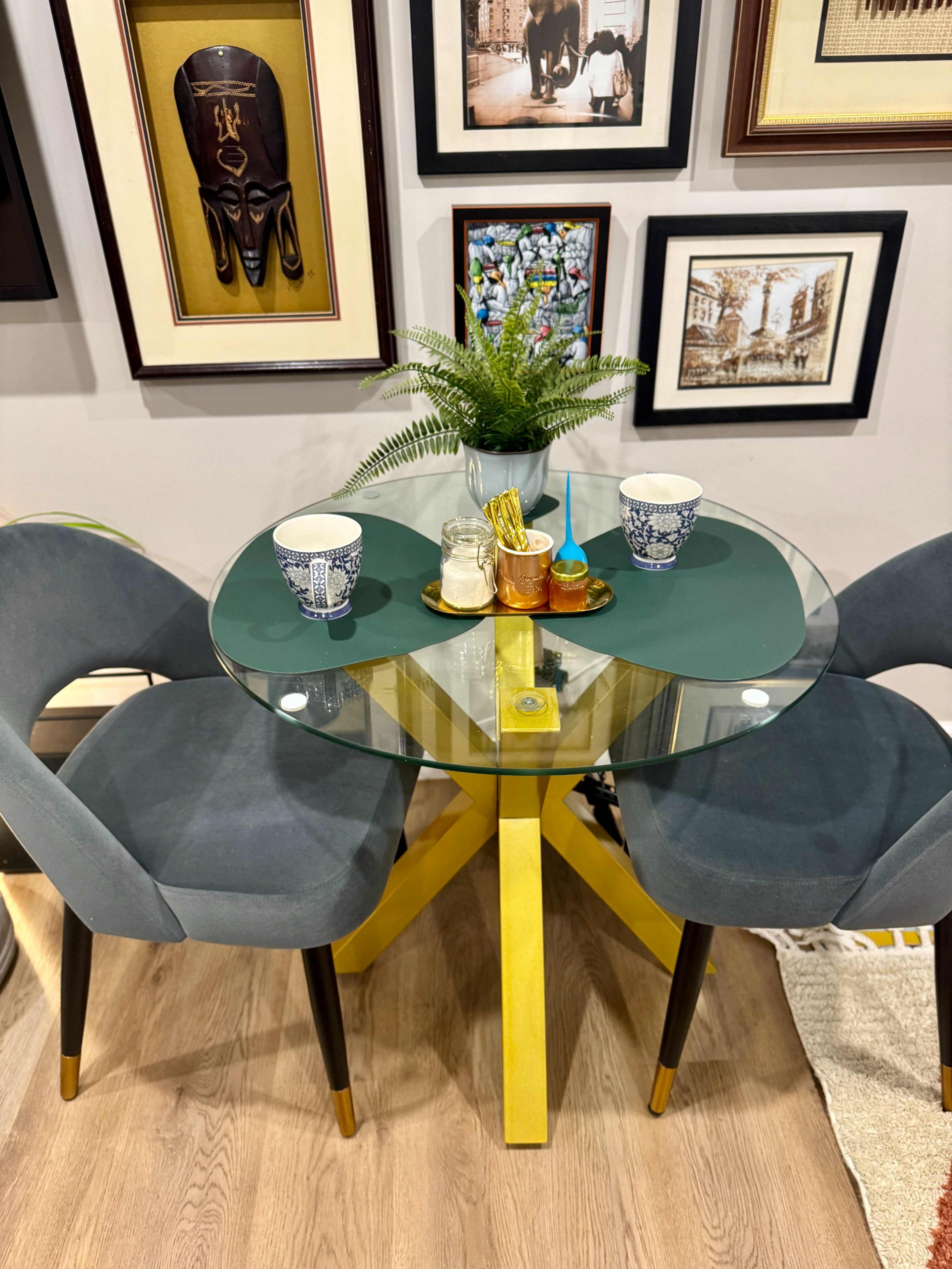 A round glass dining table with a yellow base is set between two gray chairs, adorned with decorative items and cups, and surrounded by framed art on the wall.