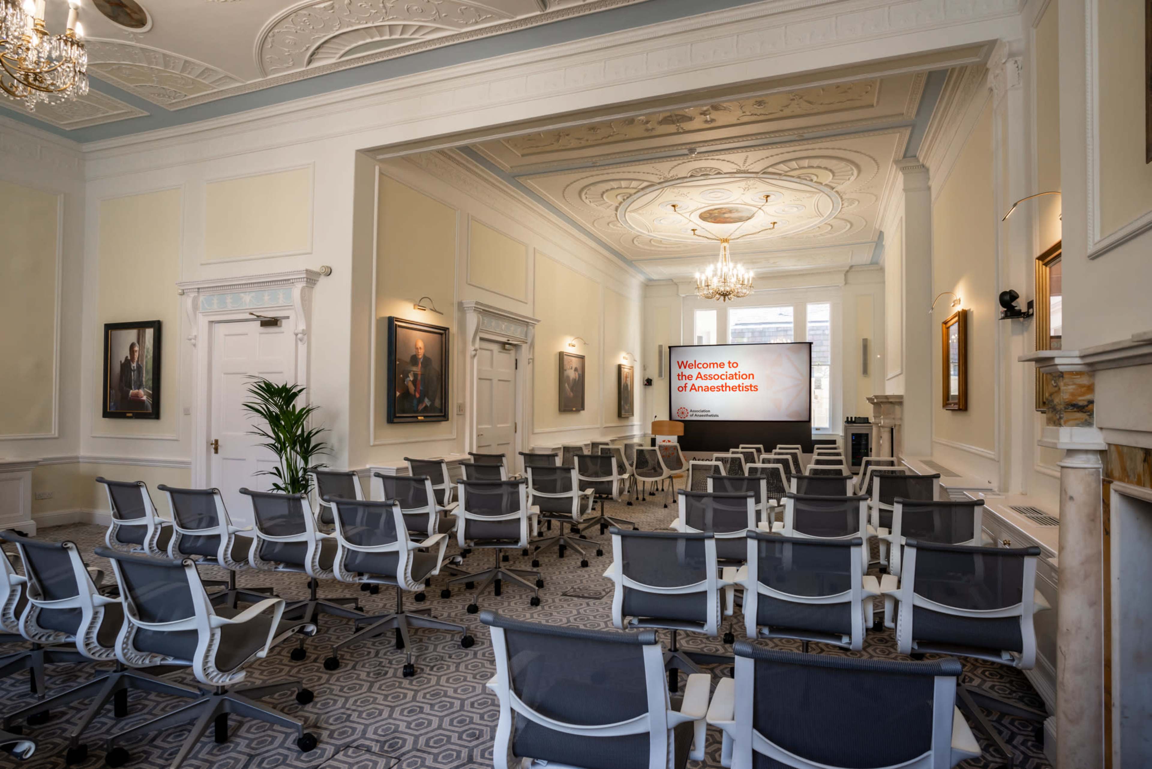 A spacious meeting room with rows of empty chairs facing a presentation screen displaying "Welcome to the Association of Anaesthetists."