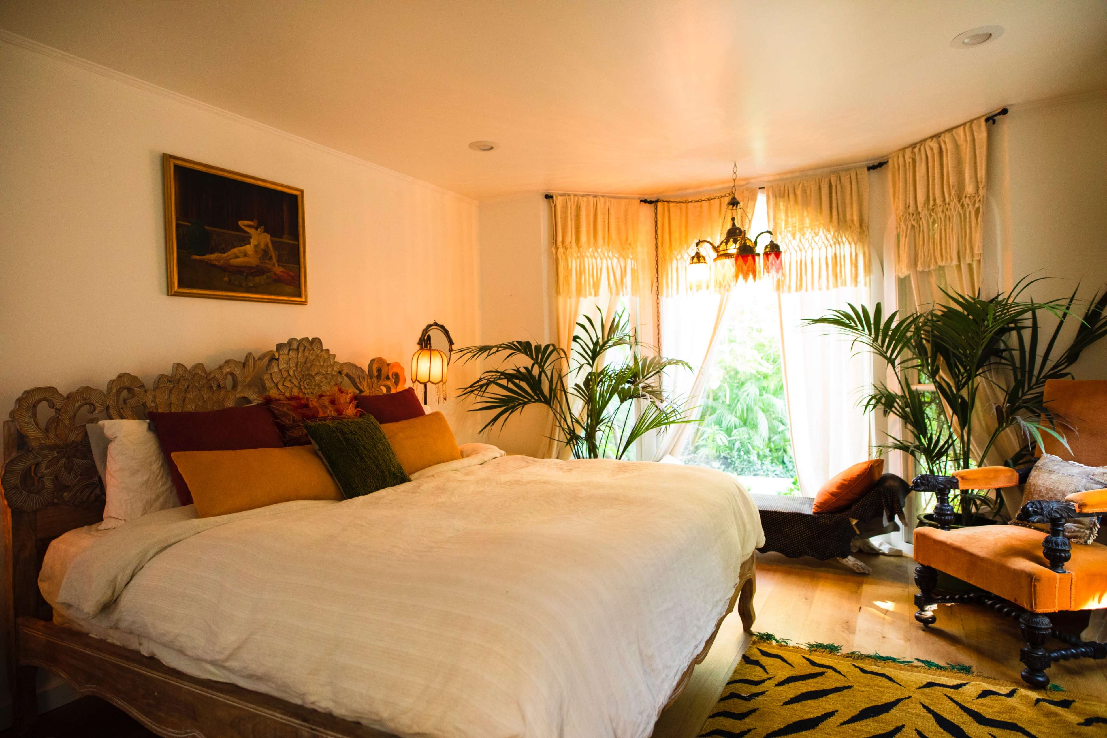A spacious bedroom featuring a large bed with decorative pillows, a window with sheer curtains, and decorative plants positioned around the room.