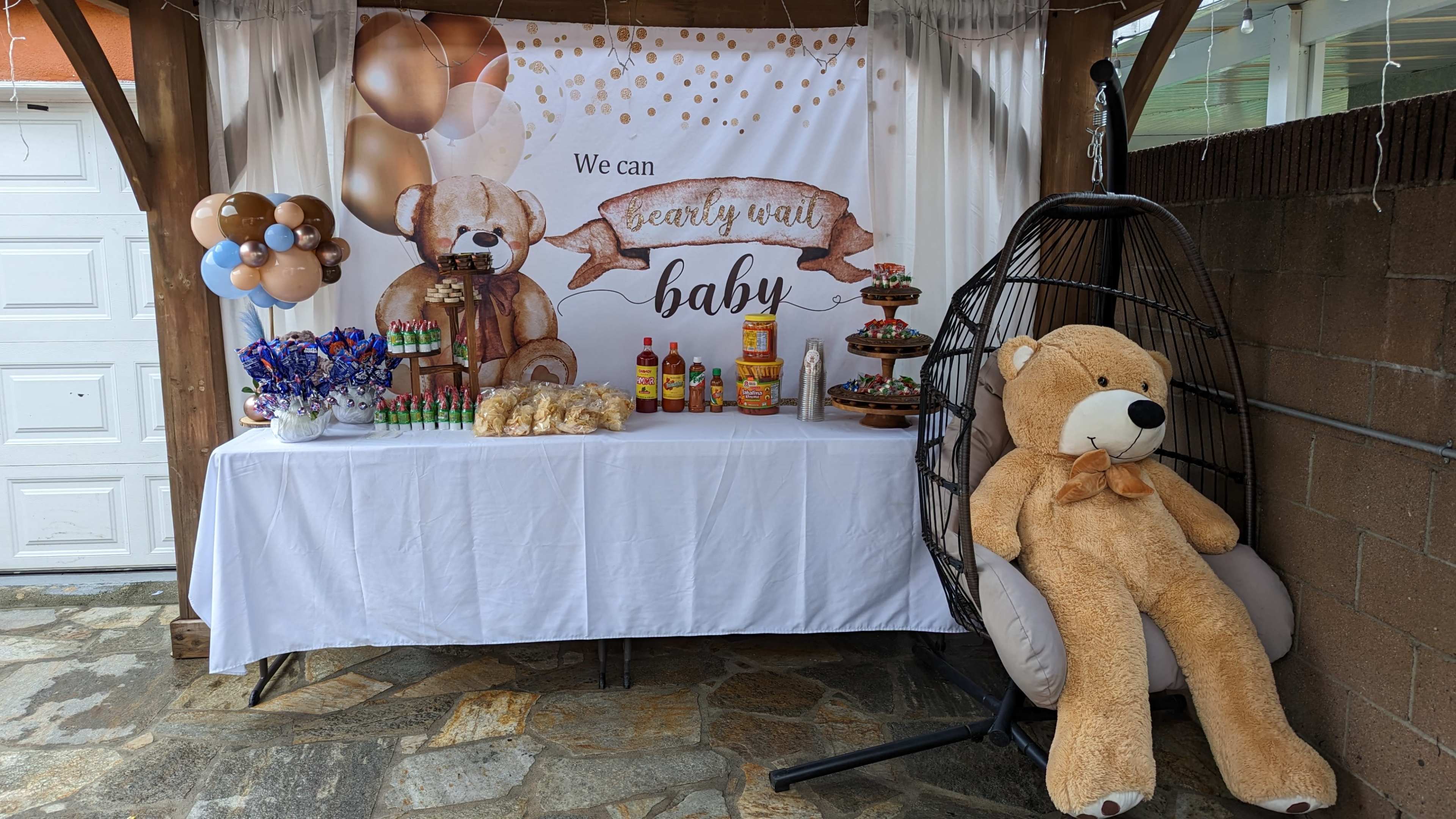 A decorated table with snacks and beverages, featuring a teddy bear theme, under a canopy with a large banner that says "We can bear-ly wait baby."