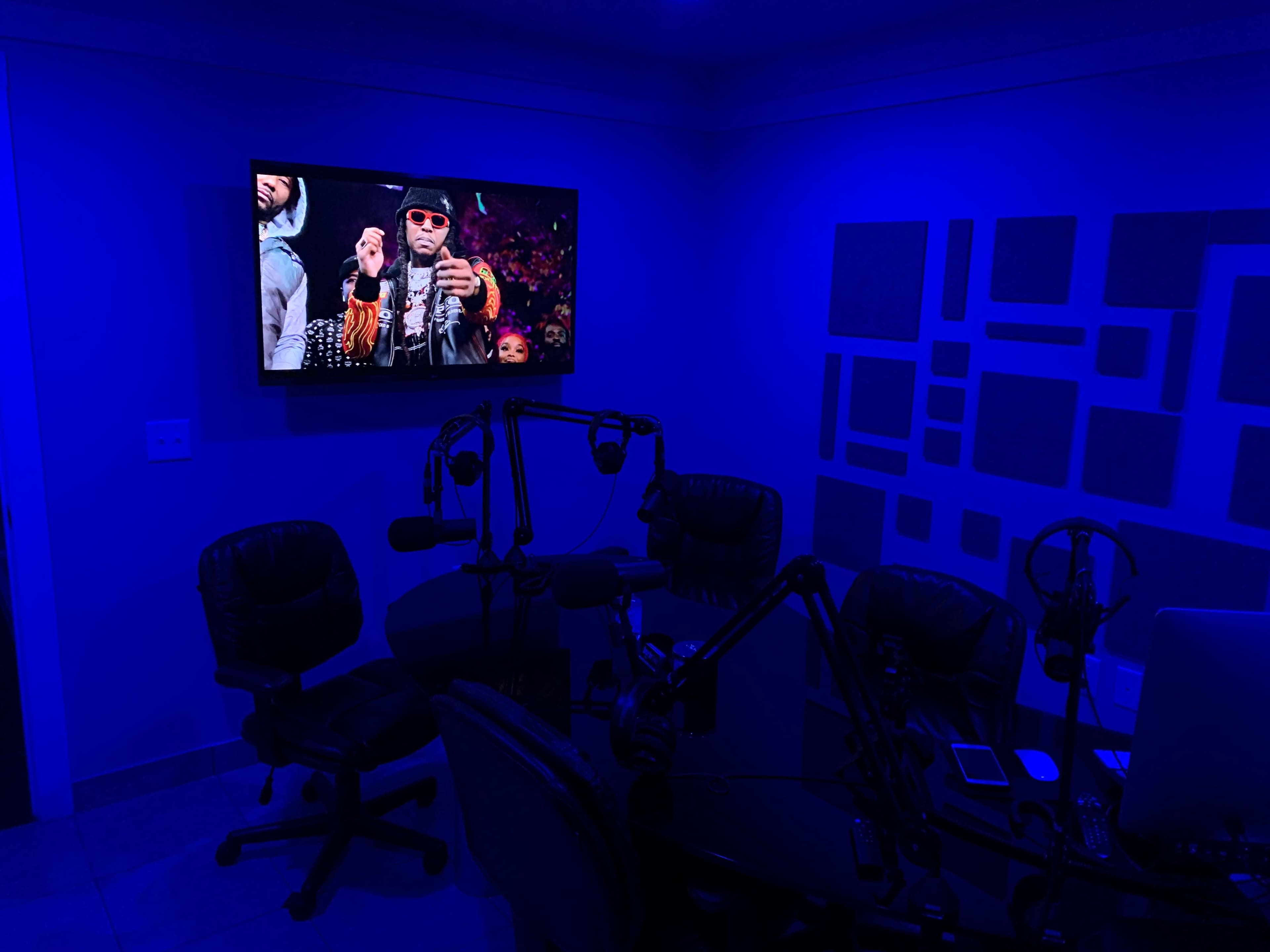 A small studio features a dark blue-lit room with multiple microphones and chairs, facing a television screen displaying a performance.