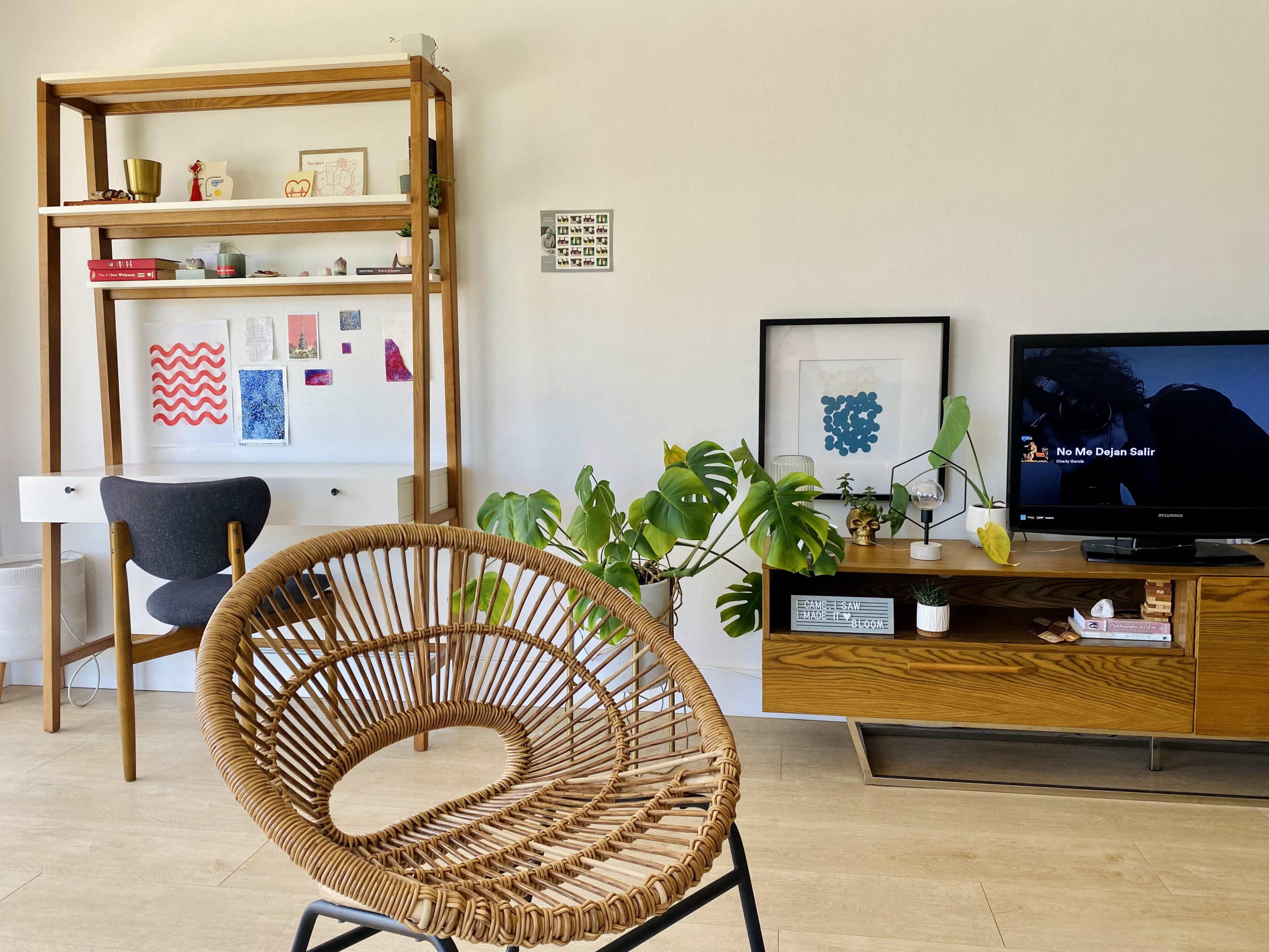 A modern living room featuring a wooden shelving unit, a framed artwork, a television on a low wooden cabinet, and a rattan chair positioned towards the camera.