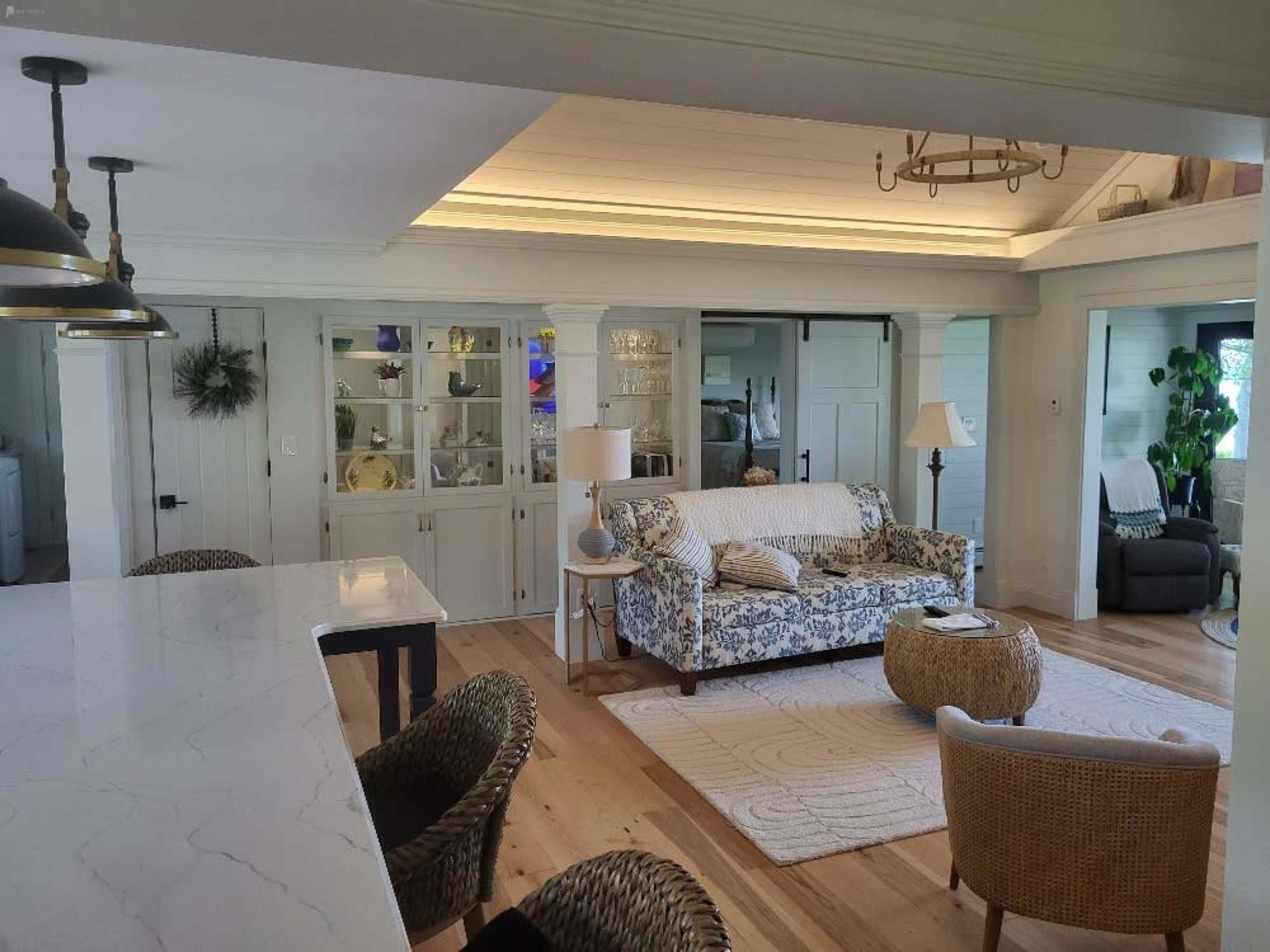 A bright living area with a floral-patterned couch, a glass-front cabinet displaying decor, and a dining table with woven chairs.