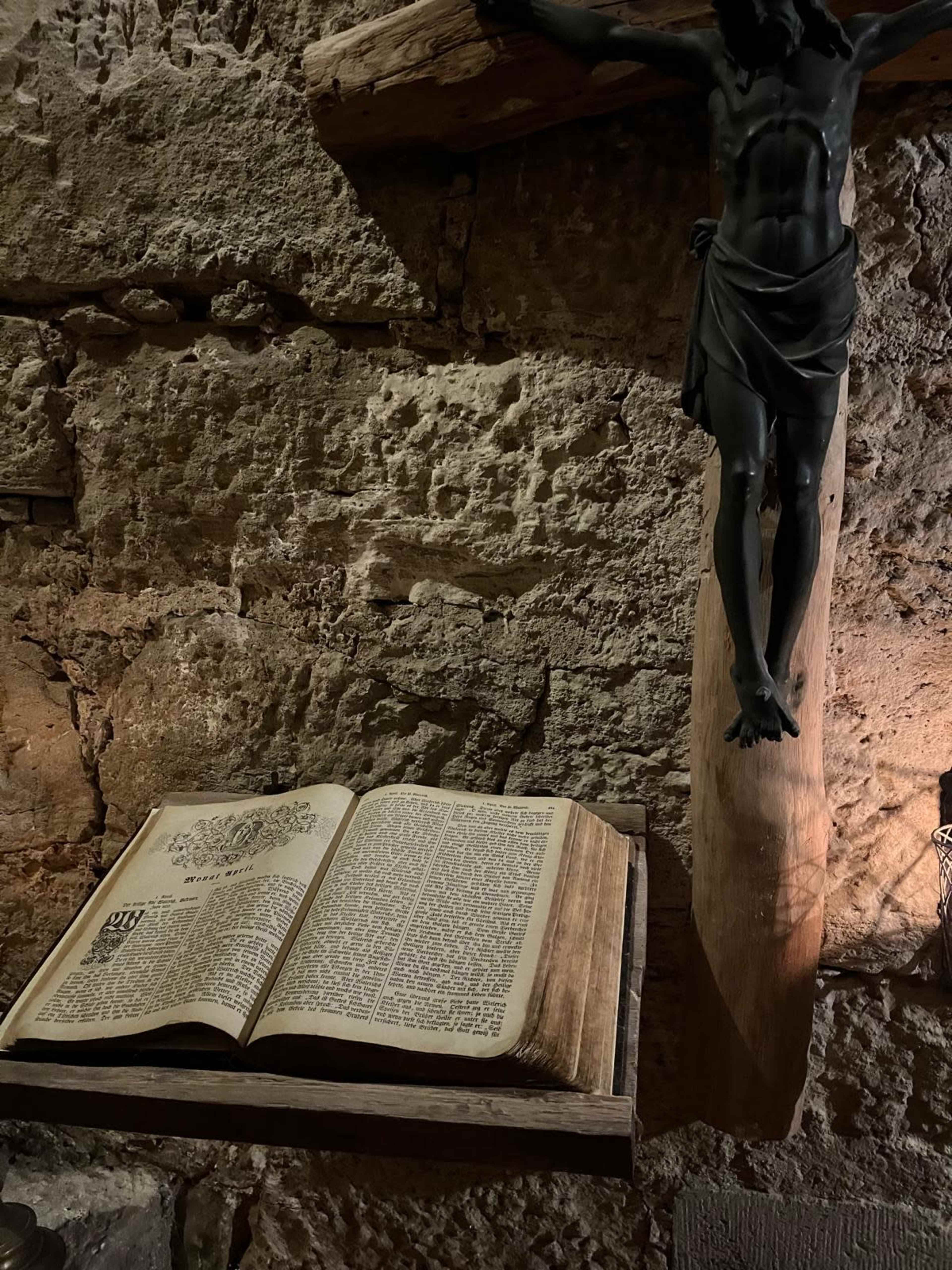 An open book is displayed on a wooden shelf next to a wooden crucifix mounted on a stone wall.