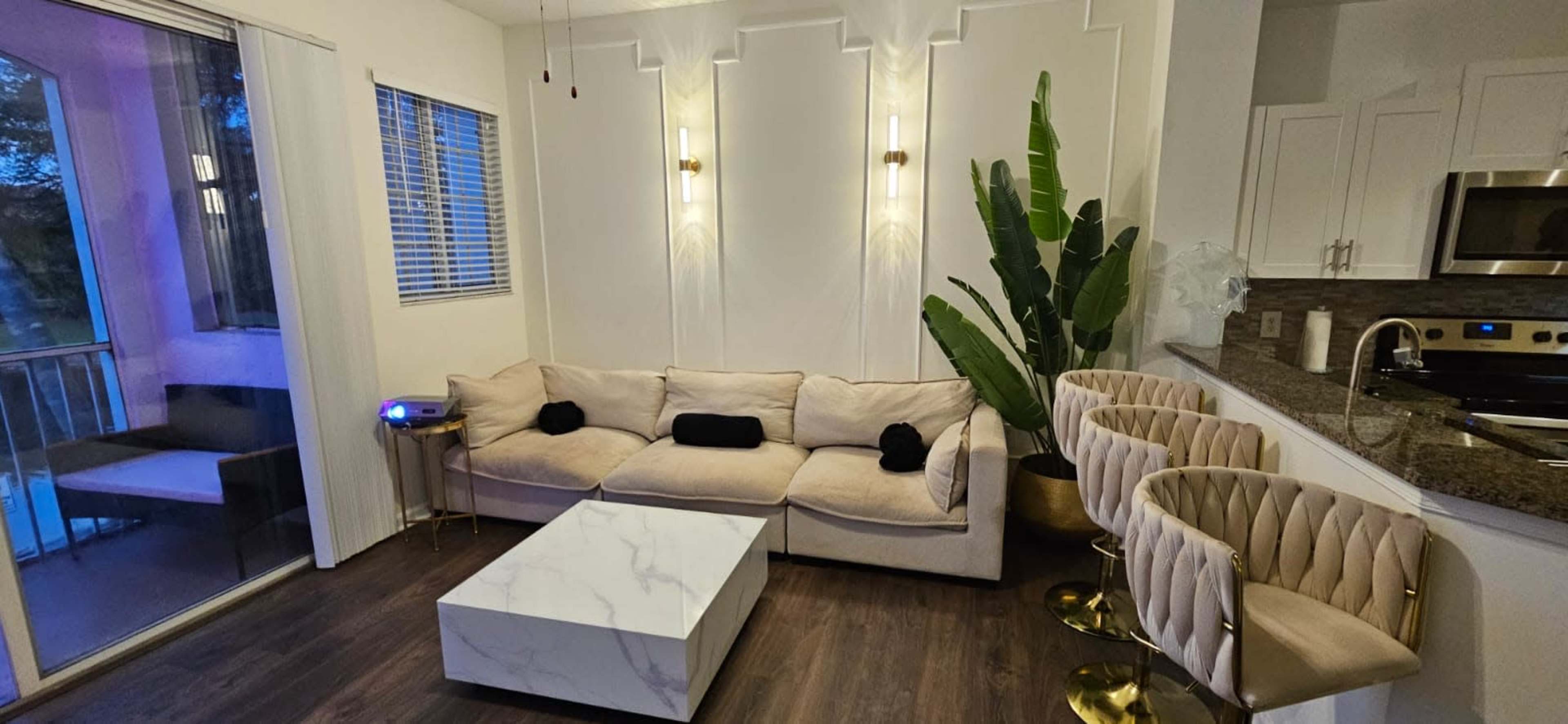 The image depicts a modern living area featuring a beige sectional sofa, a white marble coffee table, and four gold-accented bar stools near a kitchen counter.