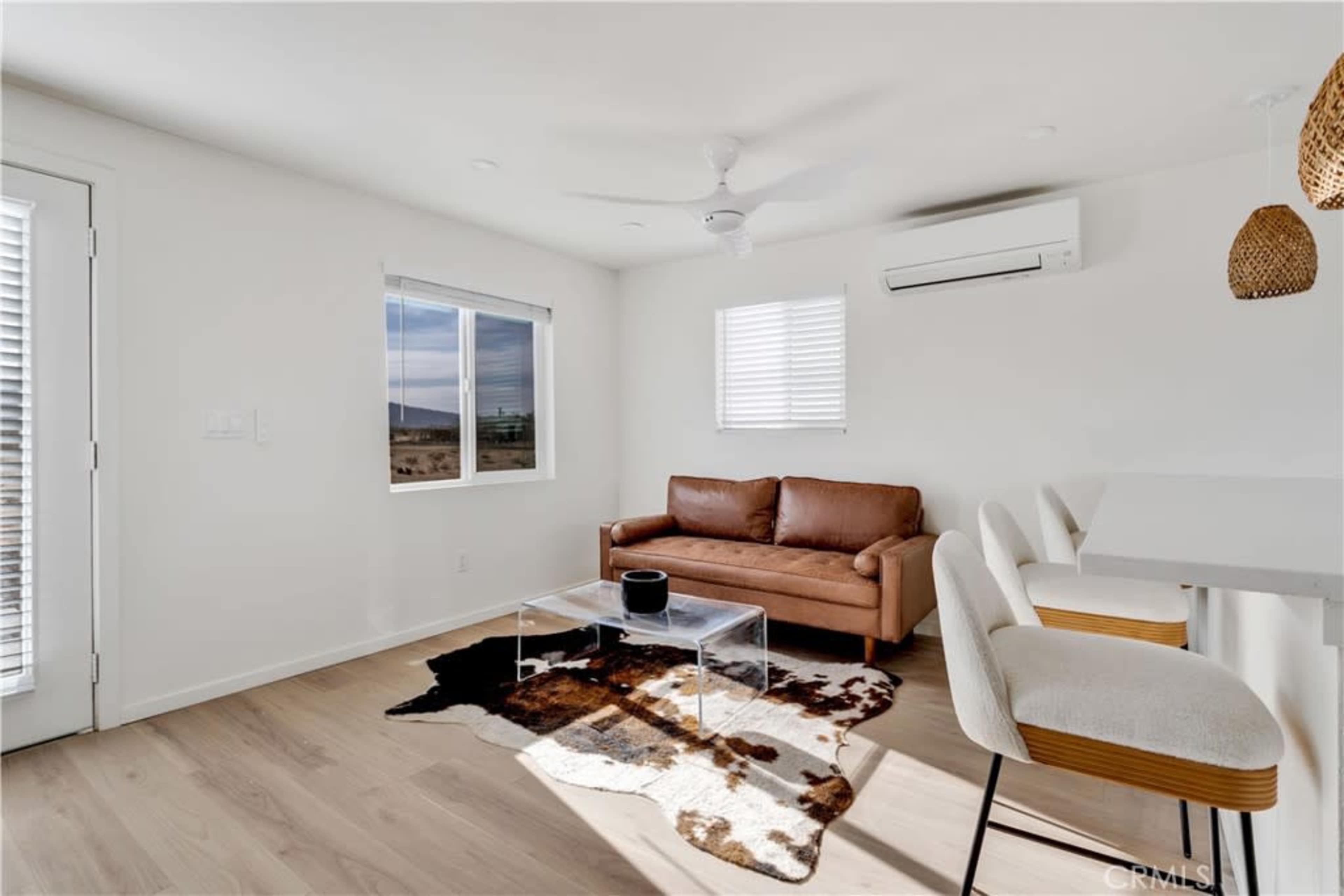 A bright, modern living room with a brown leather couch, a clear coffee table, and a small dining area featuring white stools.
