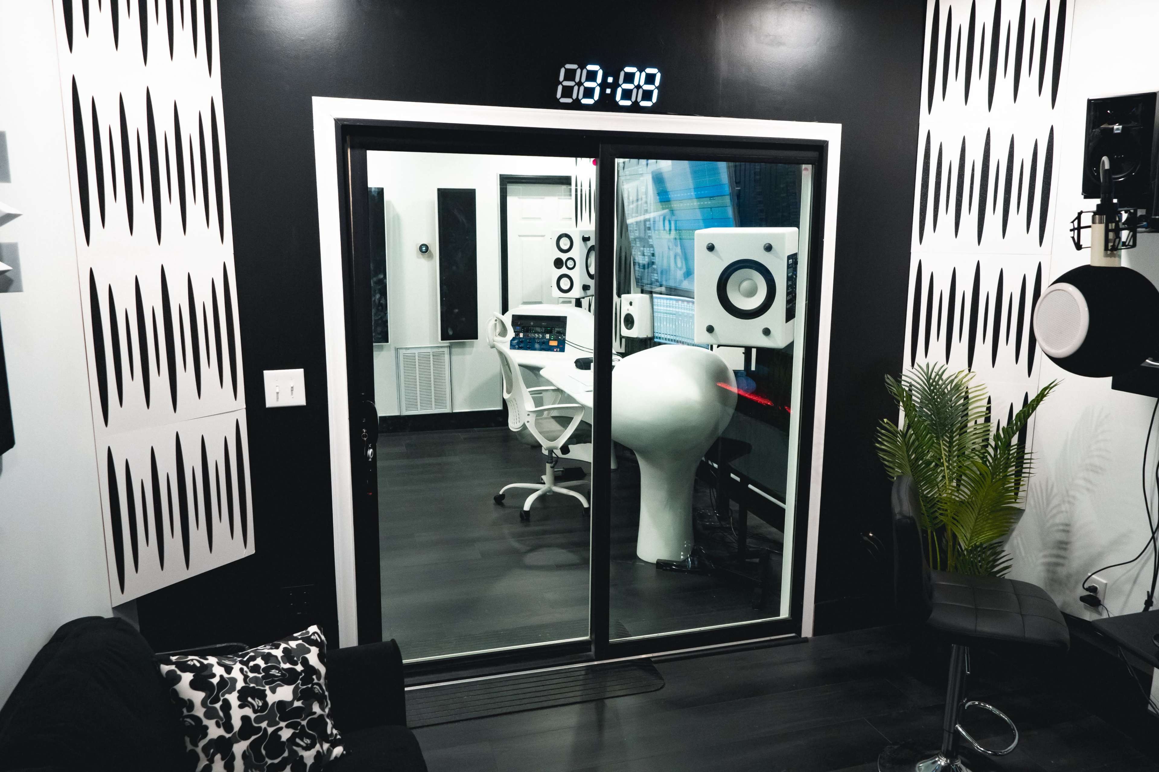 The image shows a modern recording studio with large speakers, a desk with equipment, and glass doors displaying a sleek interior.