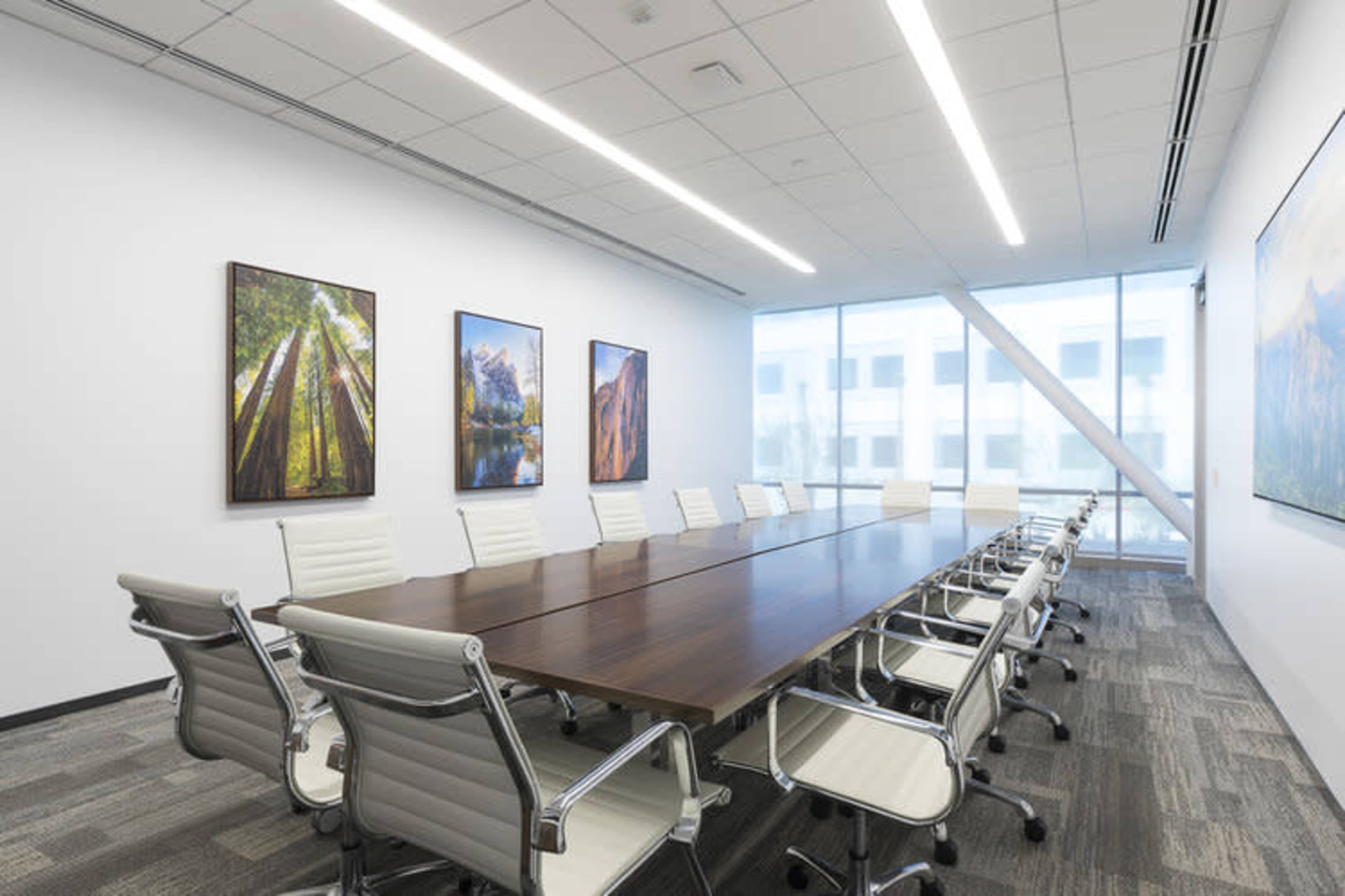 A modern conference room featuring a long wooden table surrounded by white chairs and large windows displaying artwork on the walls.