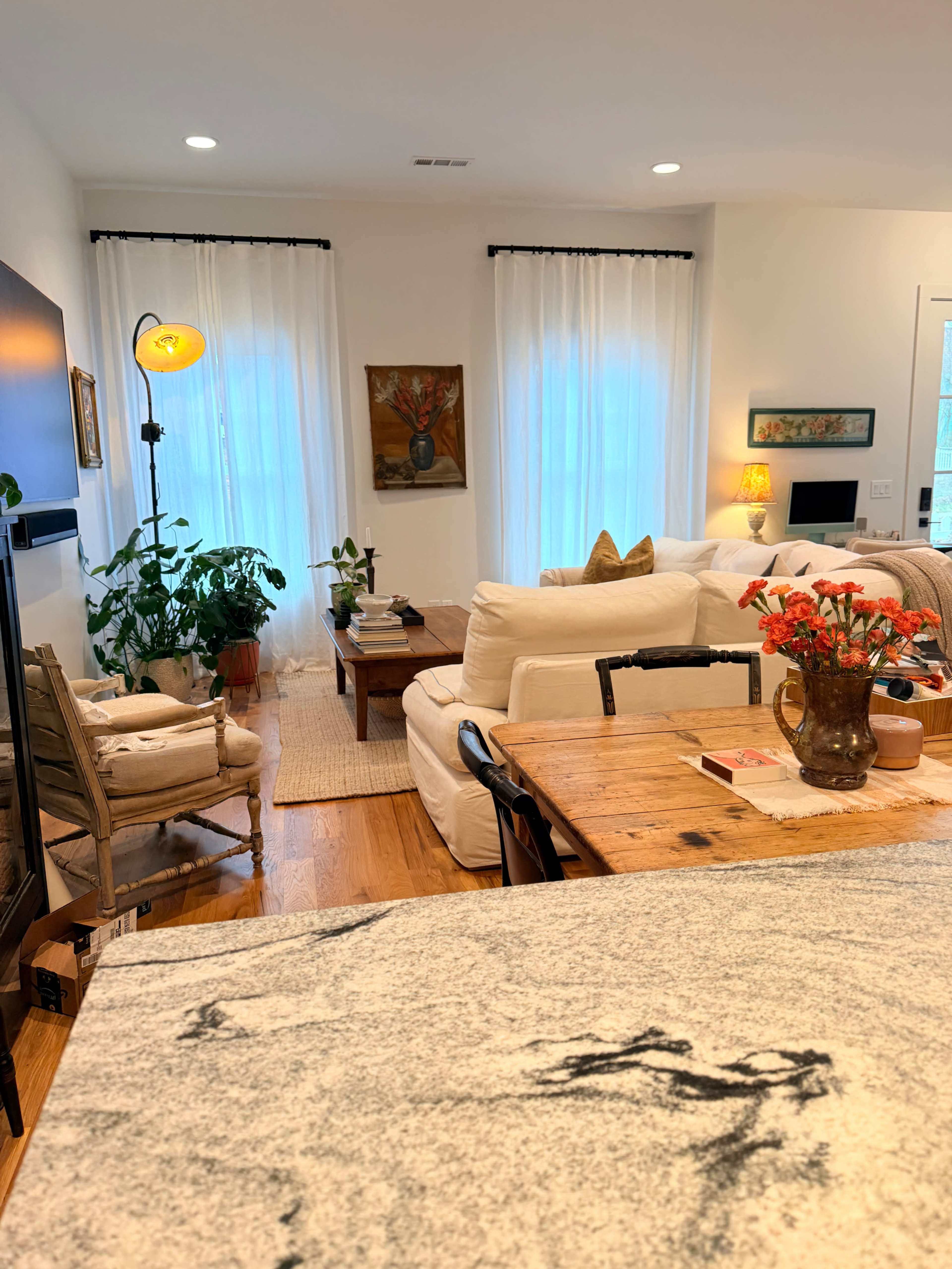 The image shows a bright living space featuring a cozy seating area with a cream-colored sofa, a wooden coffee table, and a dining table, along with plants and artwork displayed on the walls.