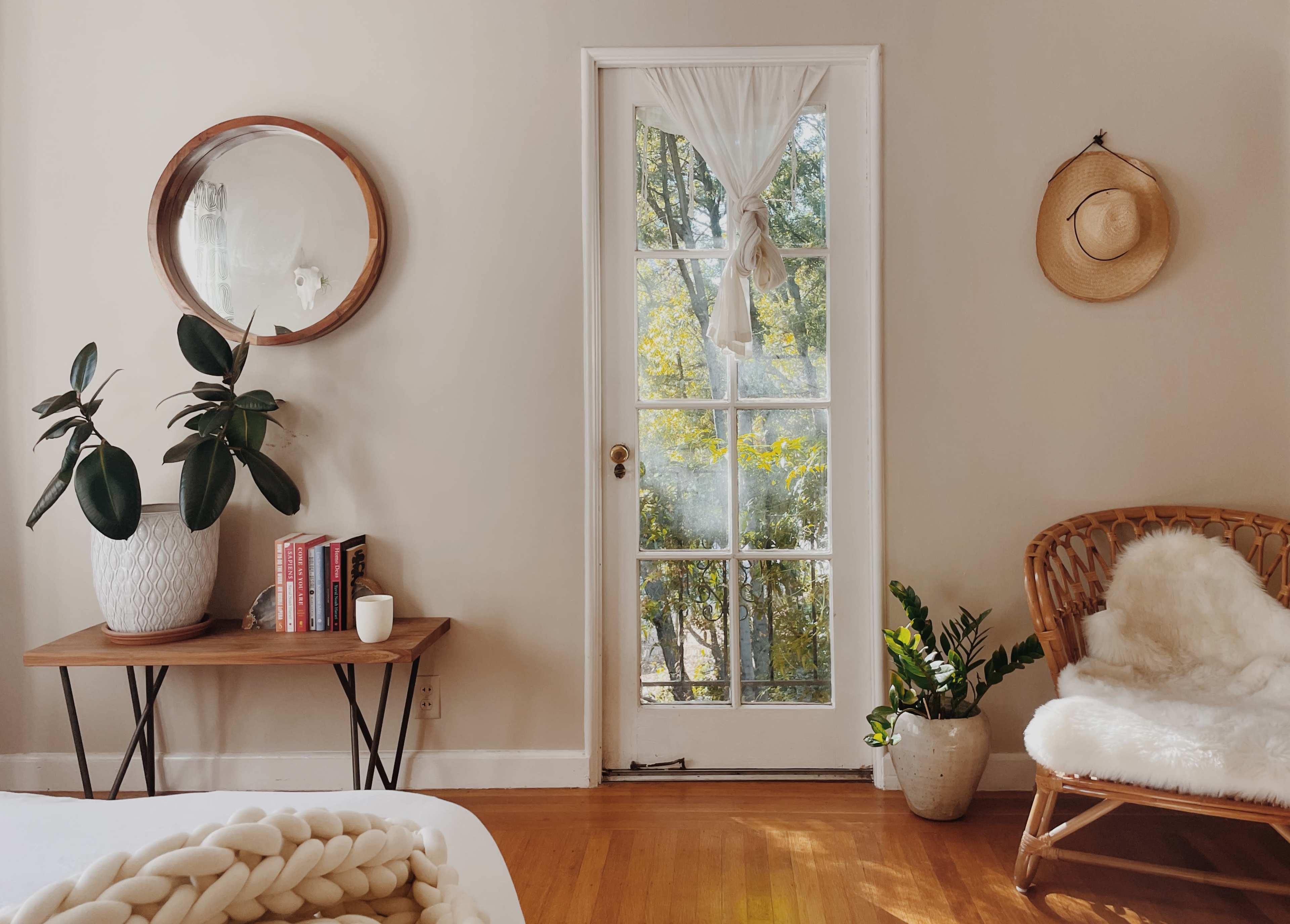 A cozy room features a wooden table with books and a candle, a wicker chair, and a glass-door entrance leading to a sunny outdoor area.