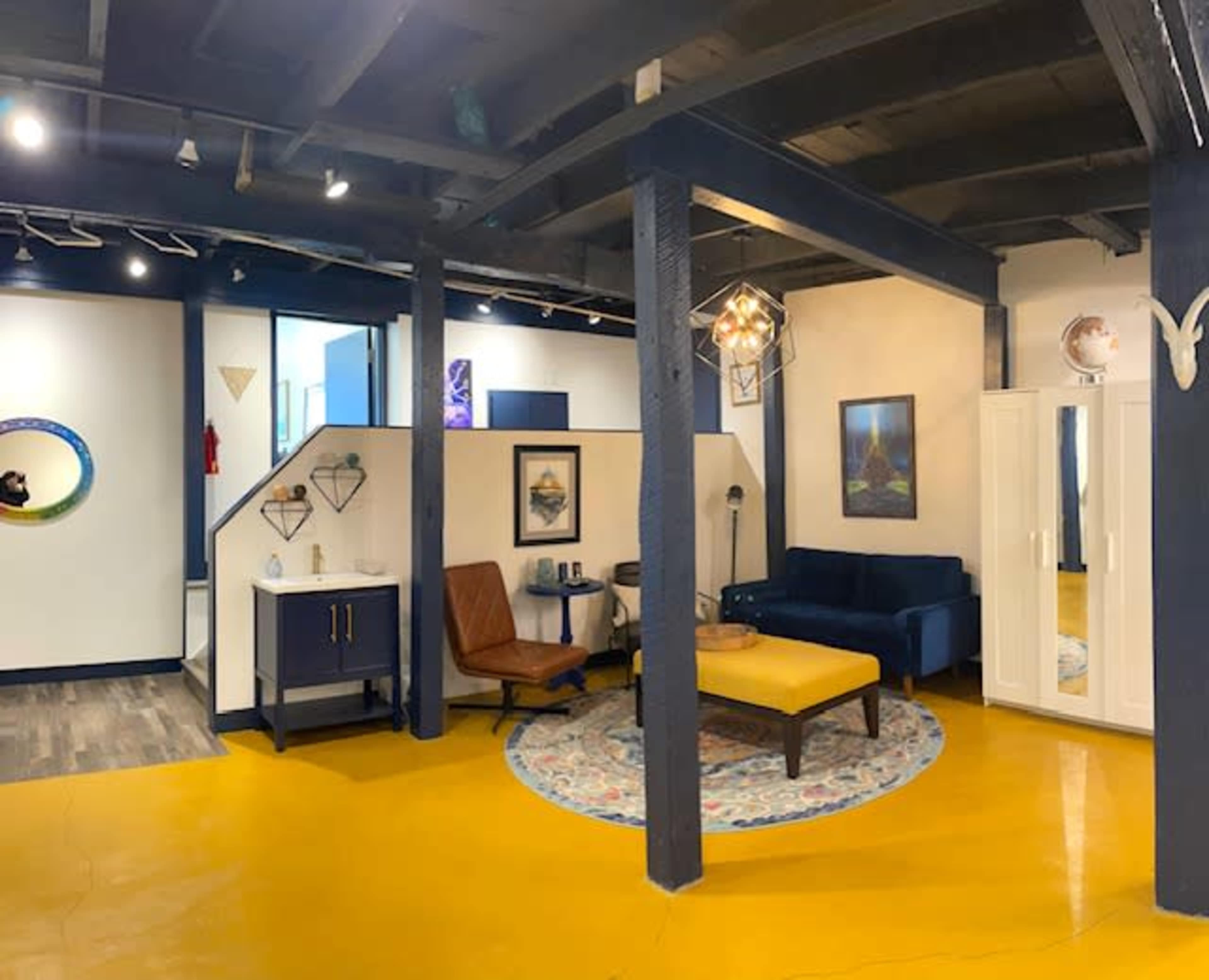 The image shows a modern interior space featuring a seating area with a blue couch, an orange armchair, and a yellow floor, complemented by decorative art on the walls.