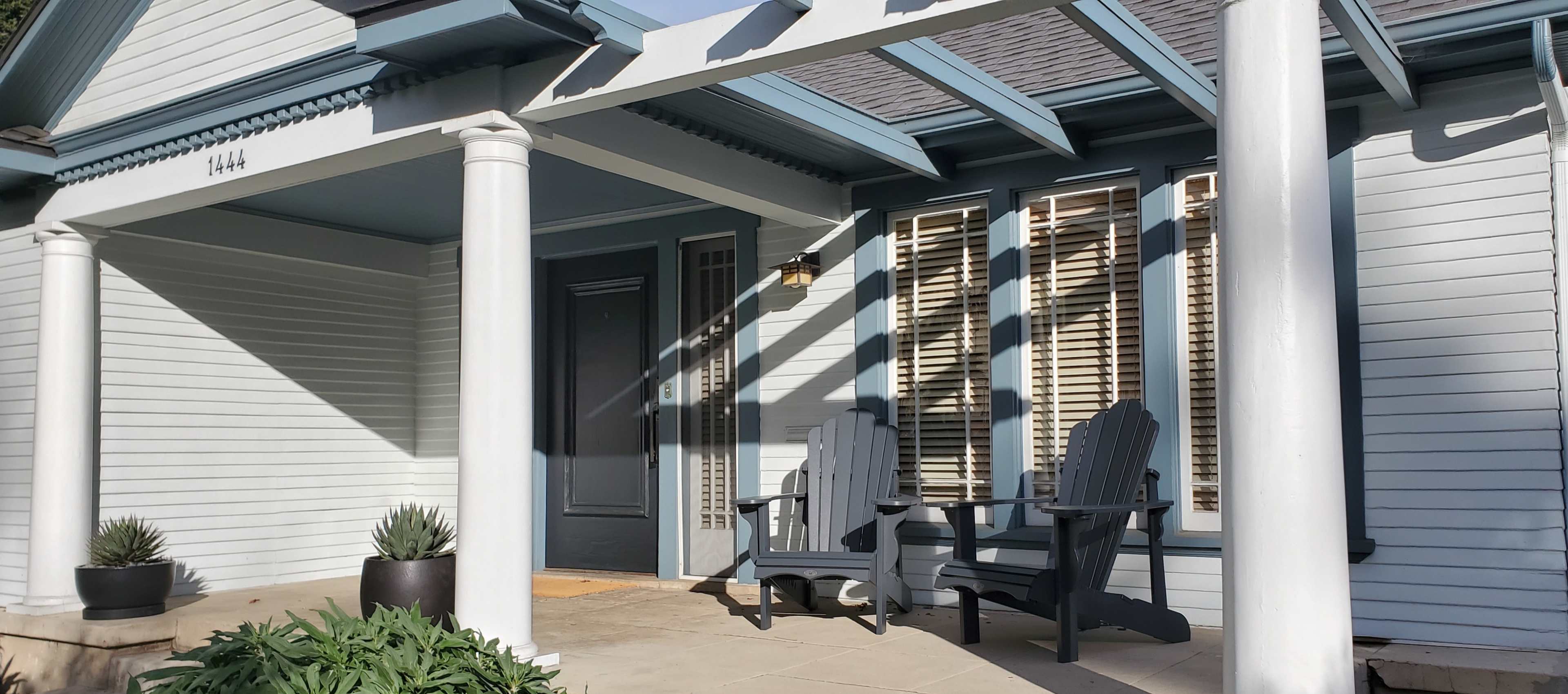 A house with a light blue exterior, featuring two gray adirondack chairs on the porch and shadow patterns cast by the columns and overhang.