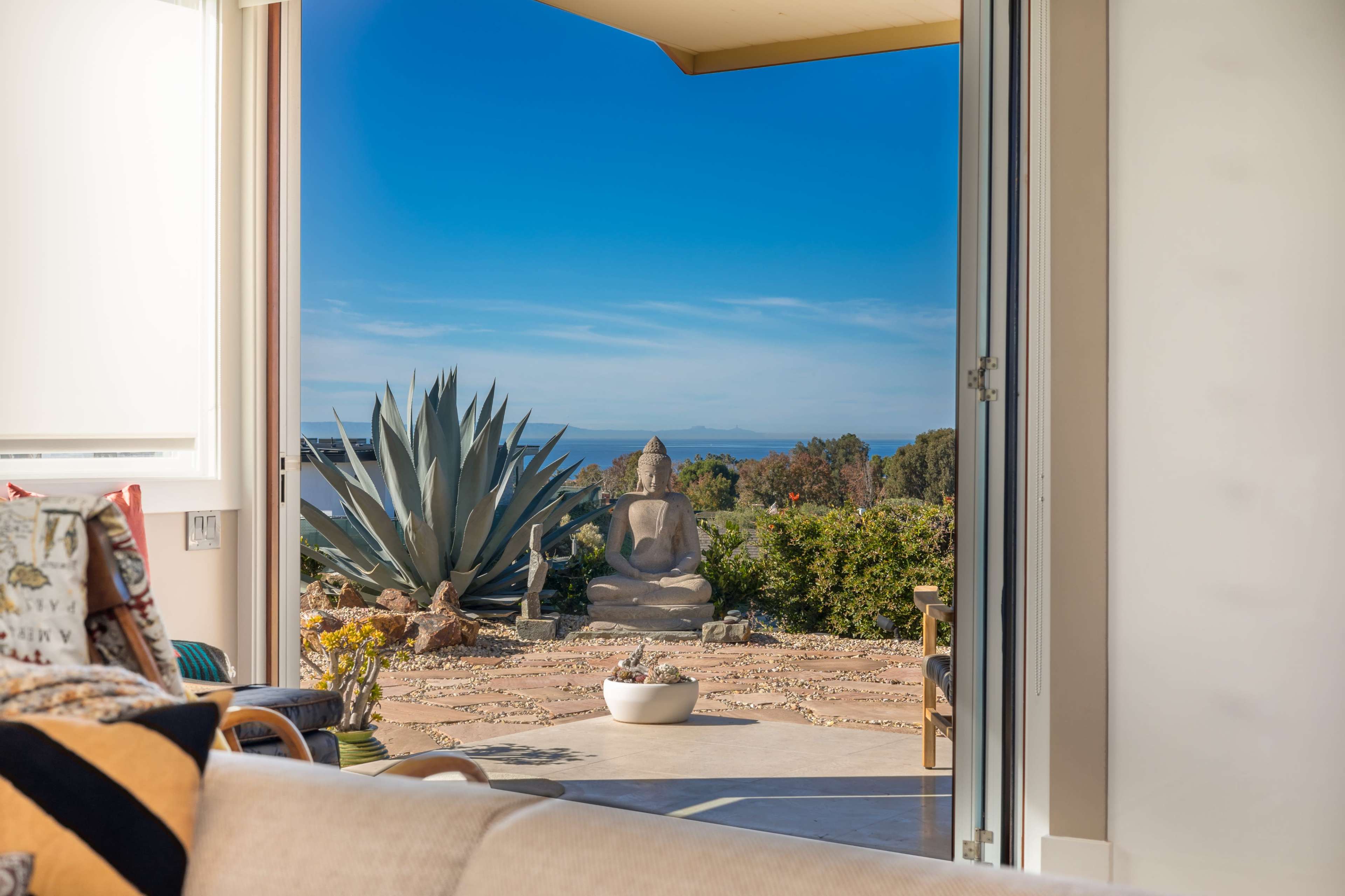 A spacious interior opens to a patio featuring a view of a large succulent and a stone Buddha statue against a clear blue sky.