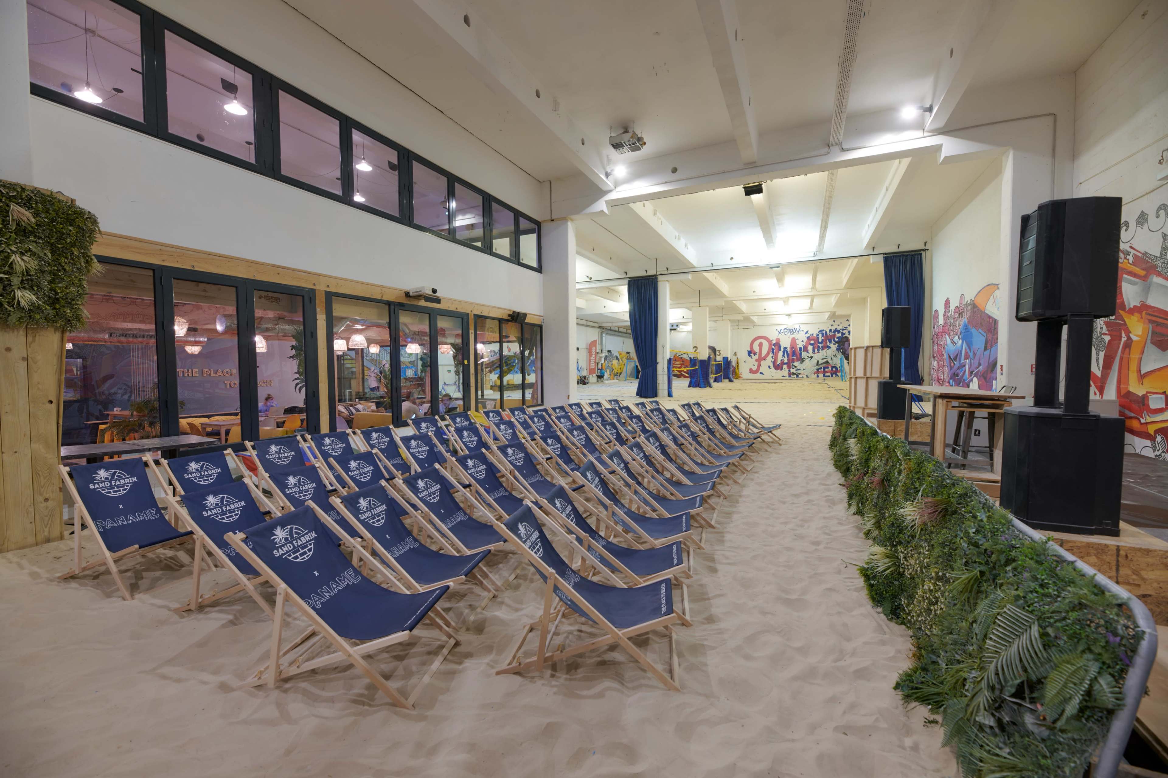 A room with chairs arranged in rows on sand, surrounded by colorful murals and large windows.