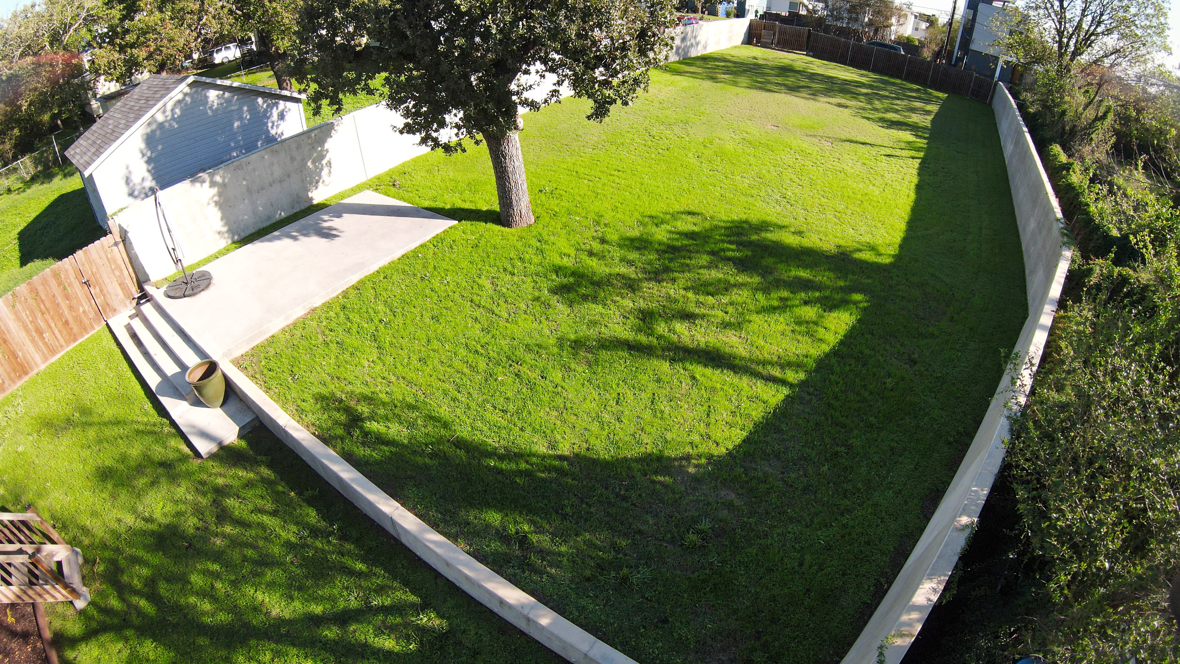 The image shows a large, grassy backyard enclosed by a concrete wall and wooden fence, with a tree casting a shadow on the lawn.