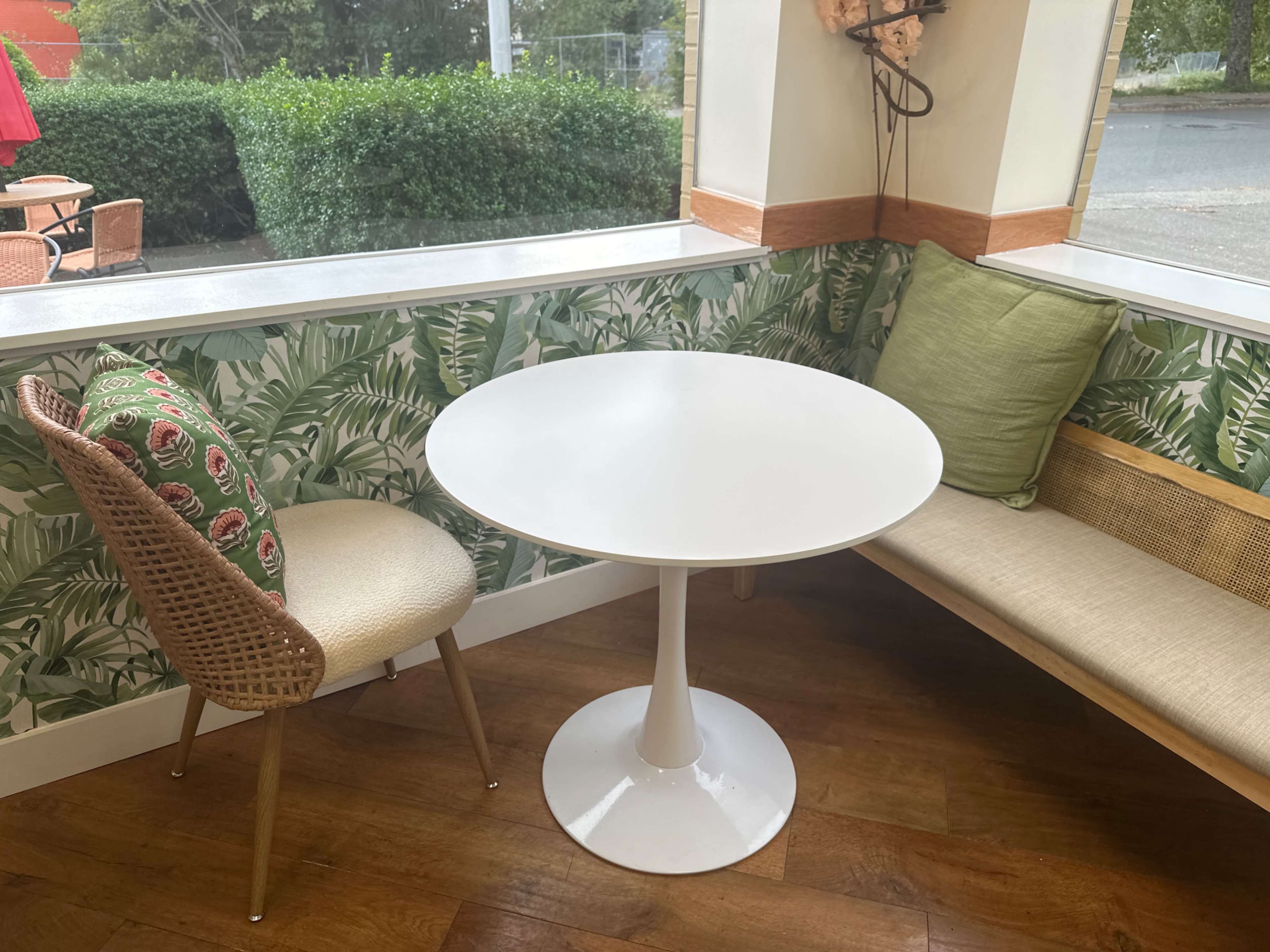A small white table is placed between a woven chair and a cushioned bench in a corner of a bright indoor space decorated with leafy wallpaper.