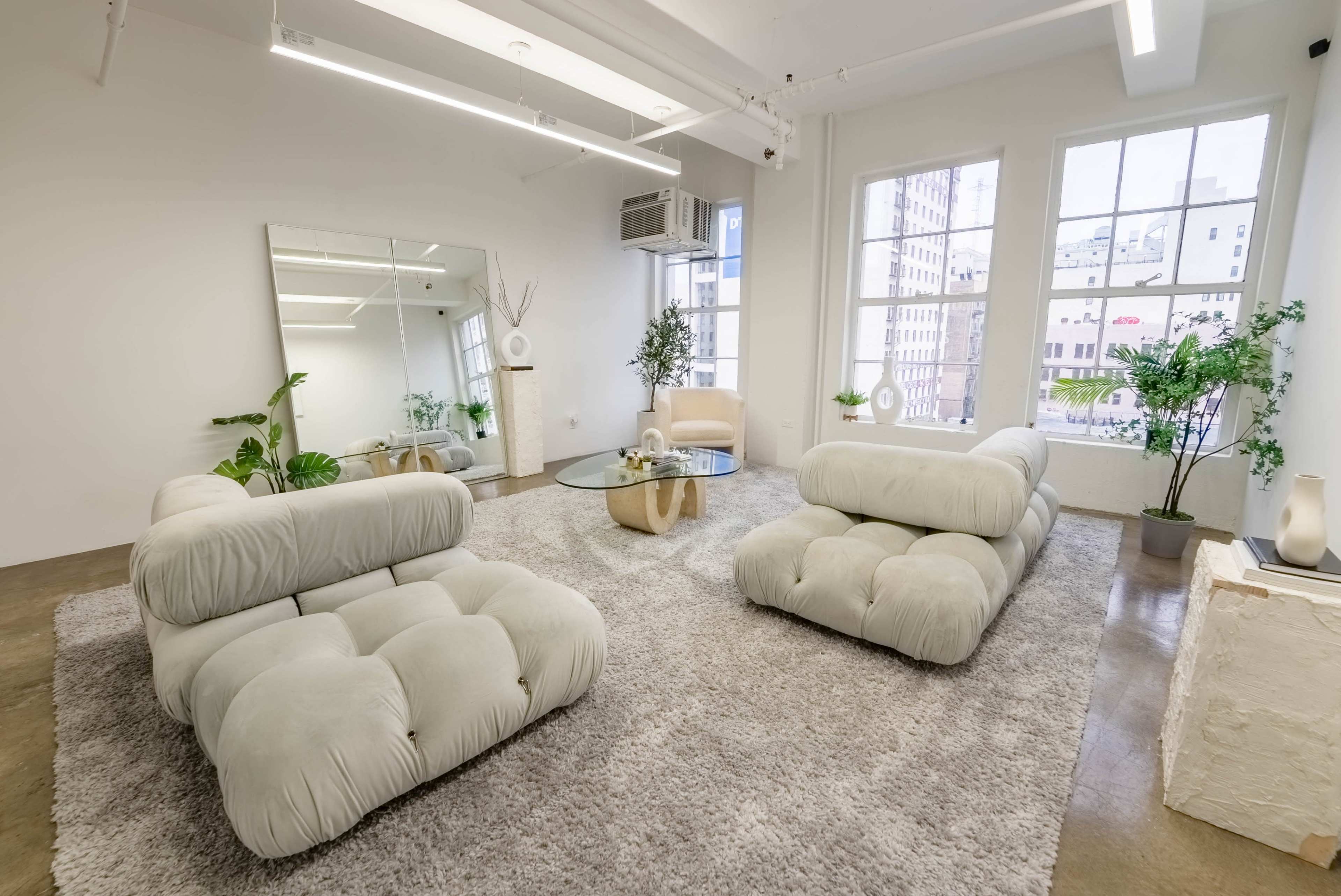 The image shows a modern, minimalist living room with two plush sofas, a glass coffee table, and large windows that allow natural light to illuminate the space.