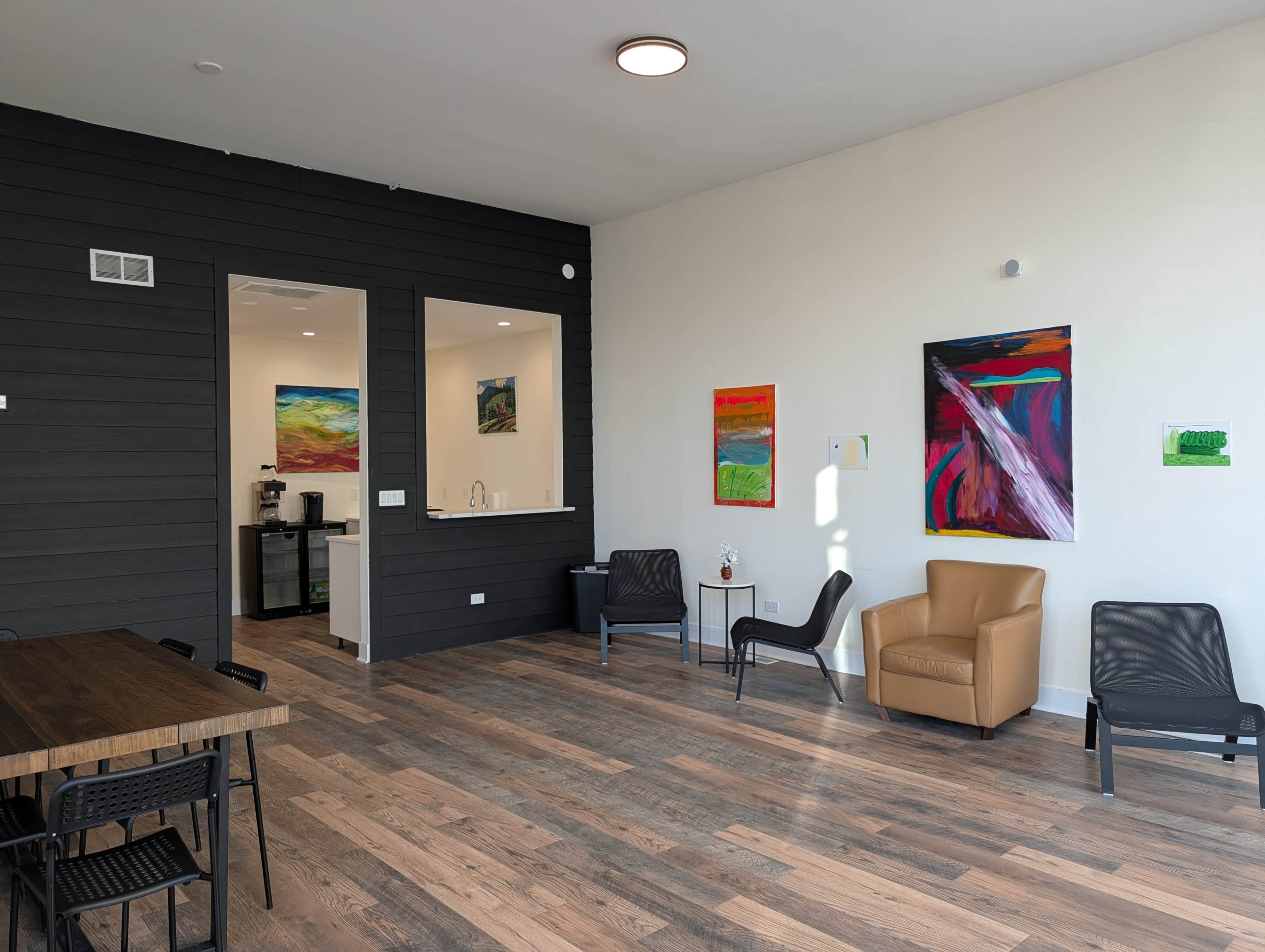 The image shows a modern interior space with a wooden floor, featuring a table and chairs on one side, and a coffee station with colorful wall art on the opposite side.
