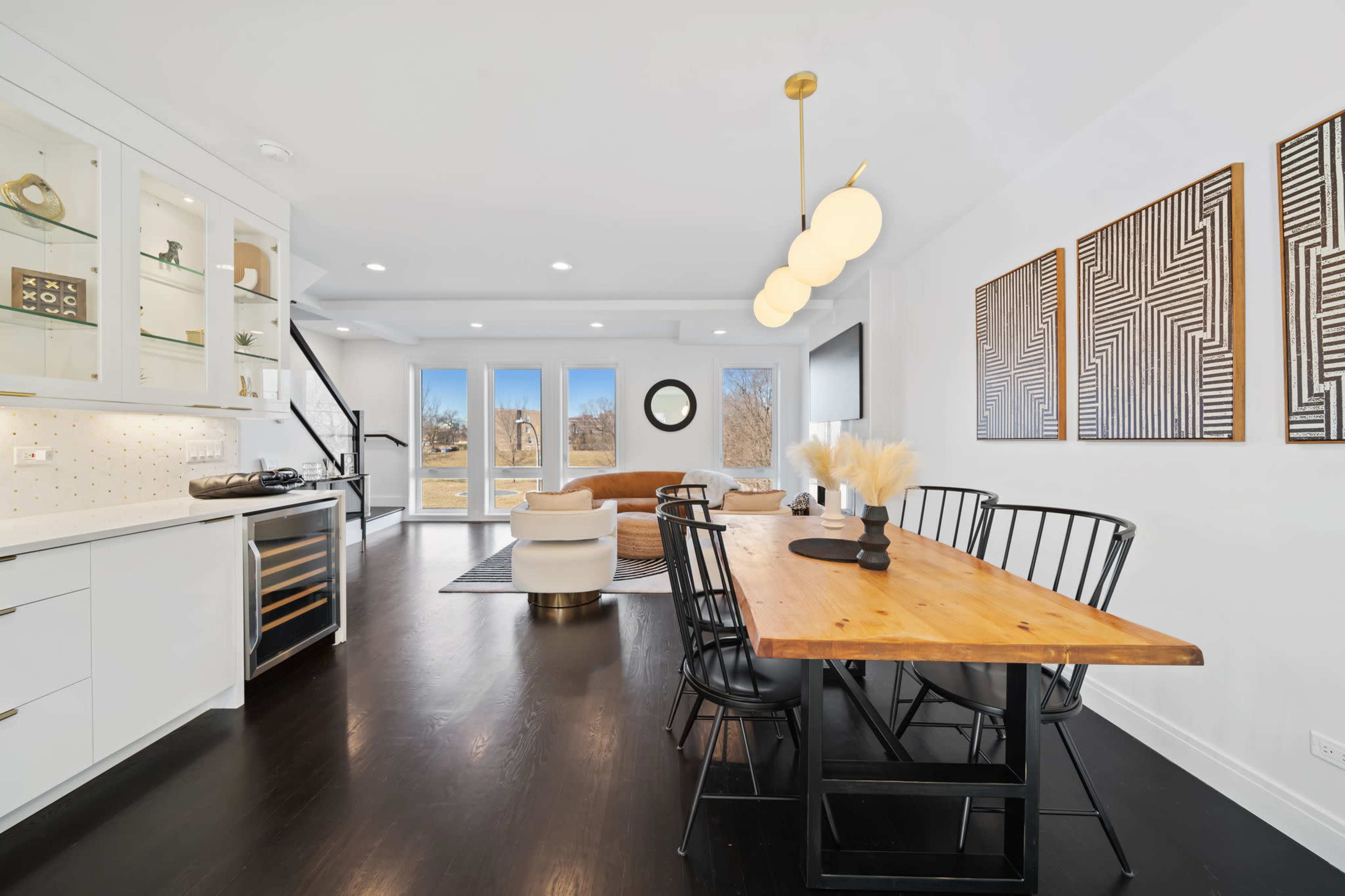 The image shows a modern dining area with a wooden table, black chairs, and a spacious living room featuring large windows and contemporary decor.